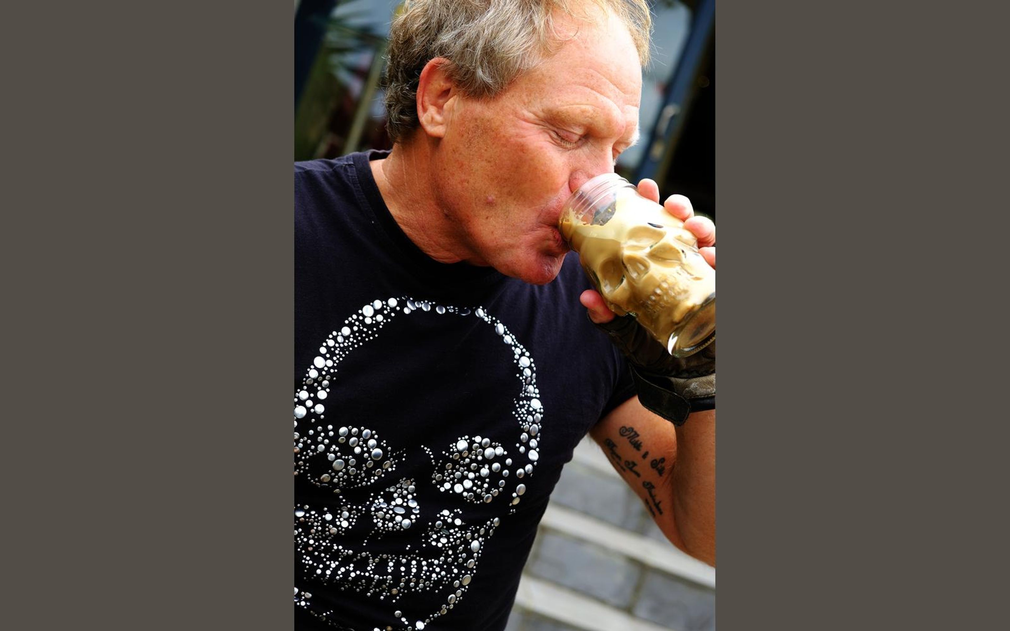 Close portrait of a man in a black skull-print shirt drinking from a gold skull-shaped glass