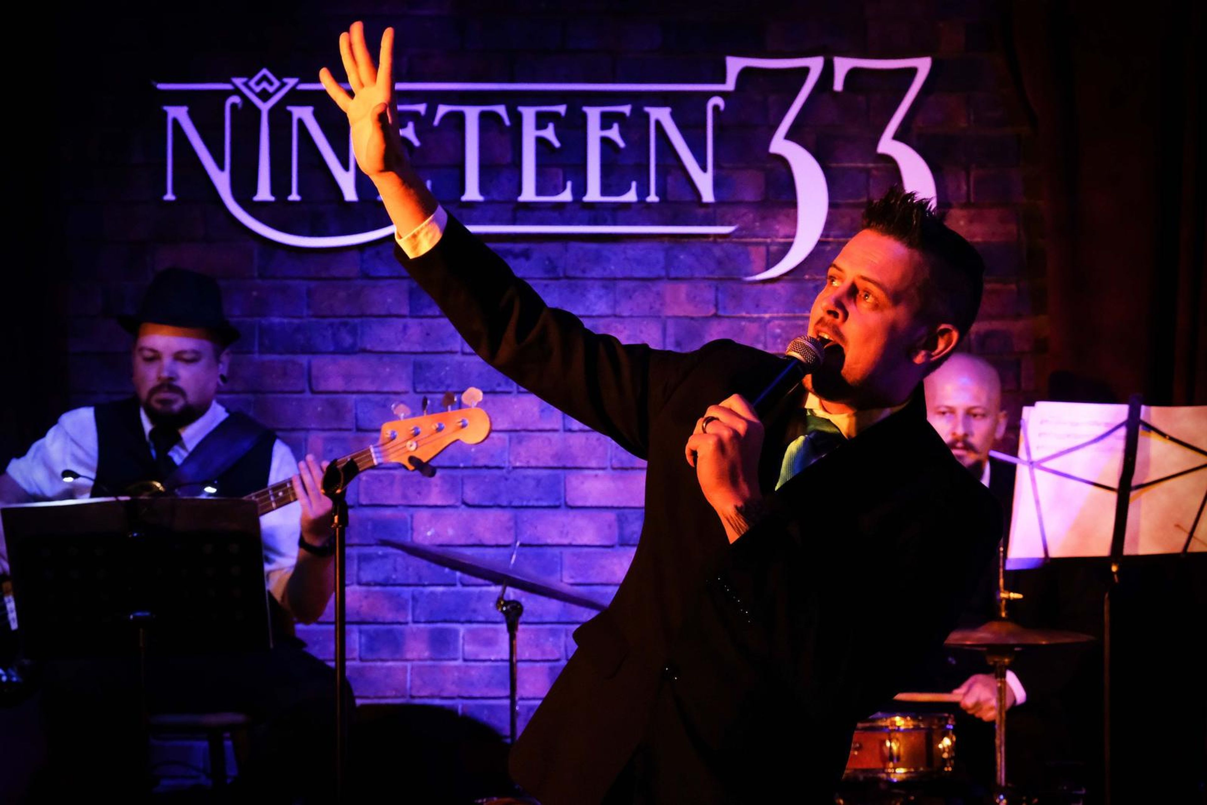 Lead singer reaching up under the Nineteen 33 neon sign with the band in purple light