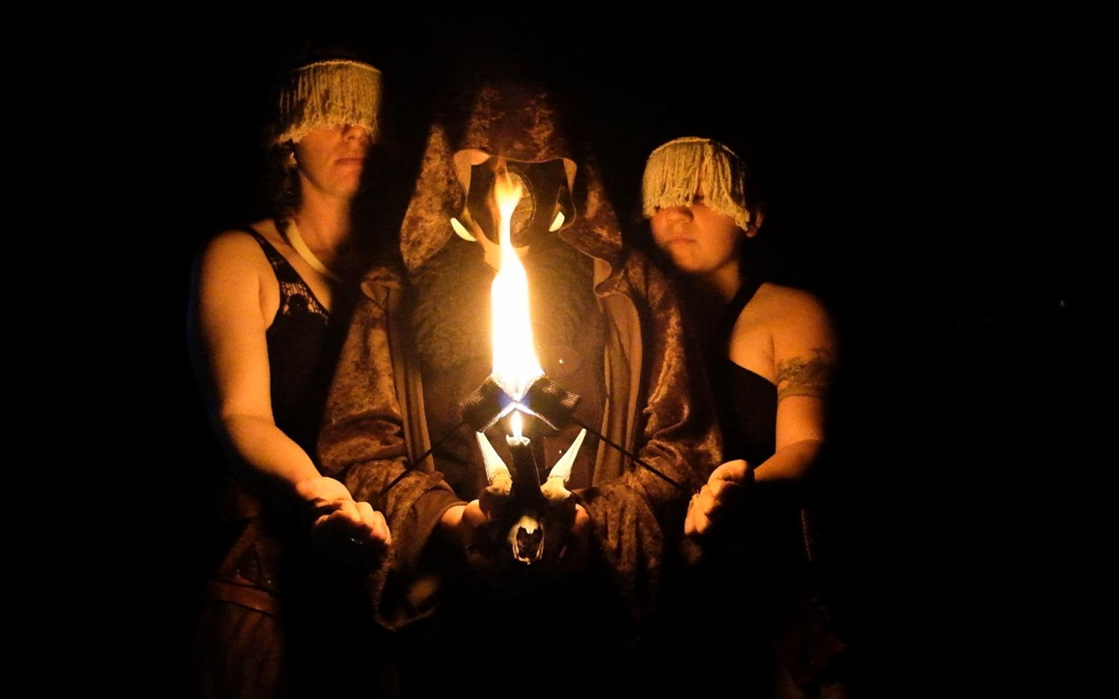 Hooded central figure holding a horned skull with a tall flame between two performers in leather and fur