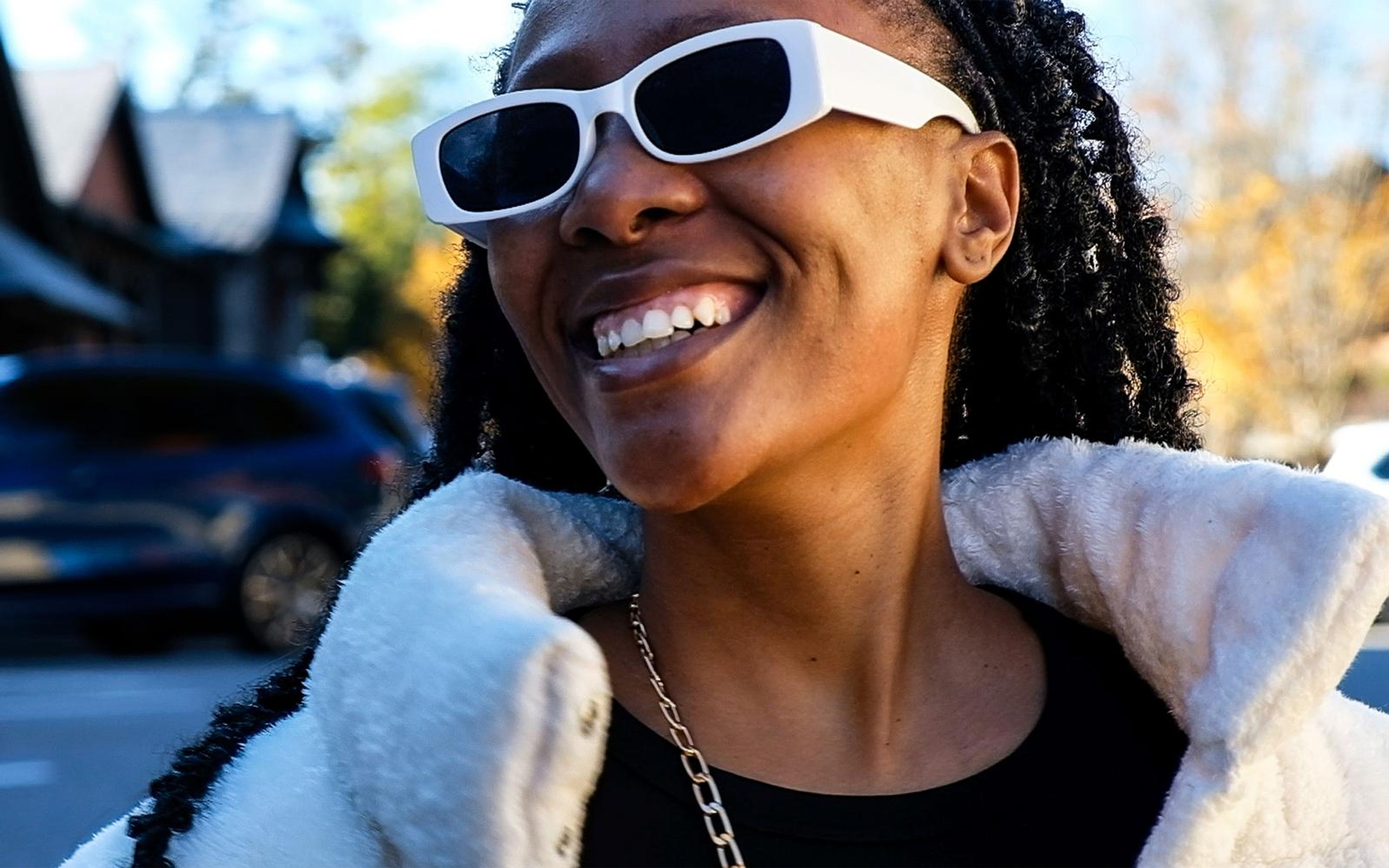 Model laughing with head tipped back, white sunglasses, fur collar, parked cars and autumn foliage behind