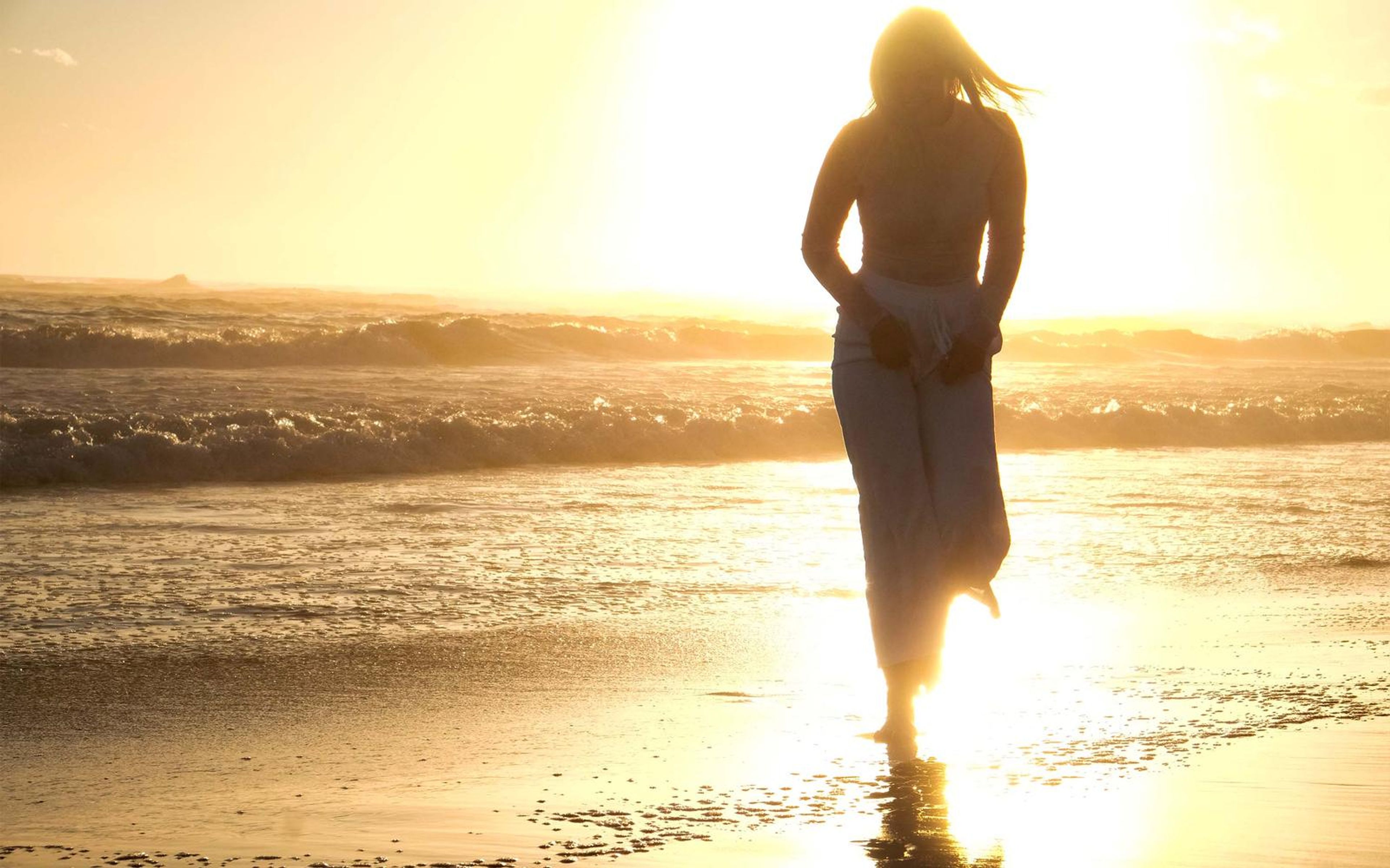 Woman standing at the shoreline holding a cloth at her waist, sun low behind her