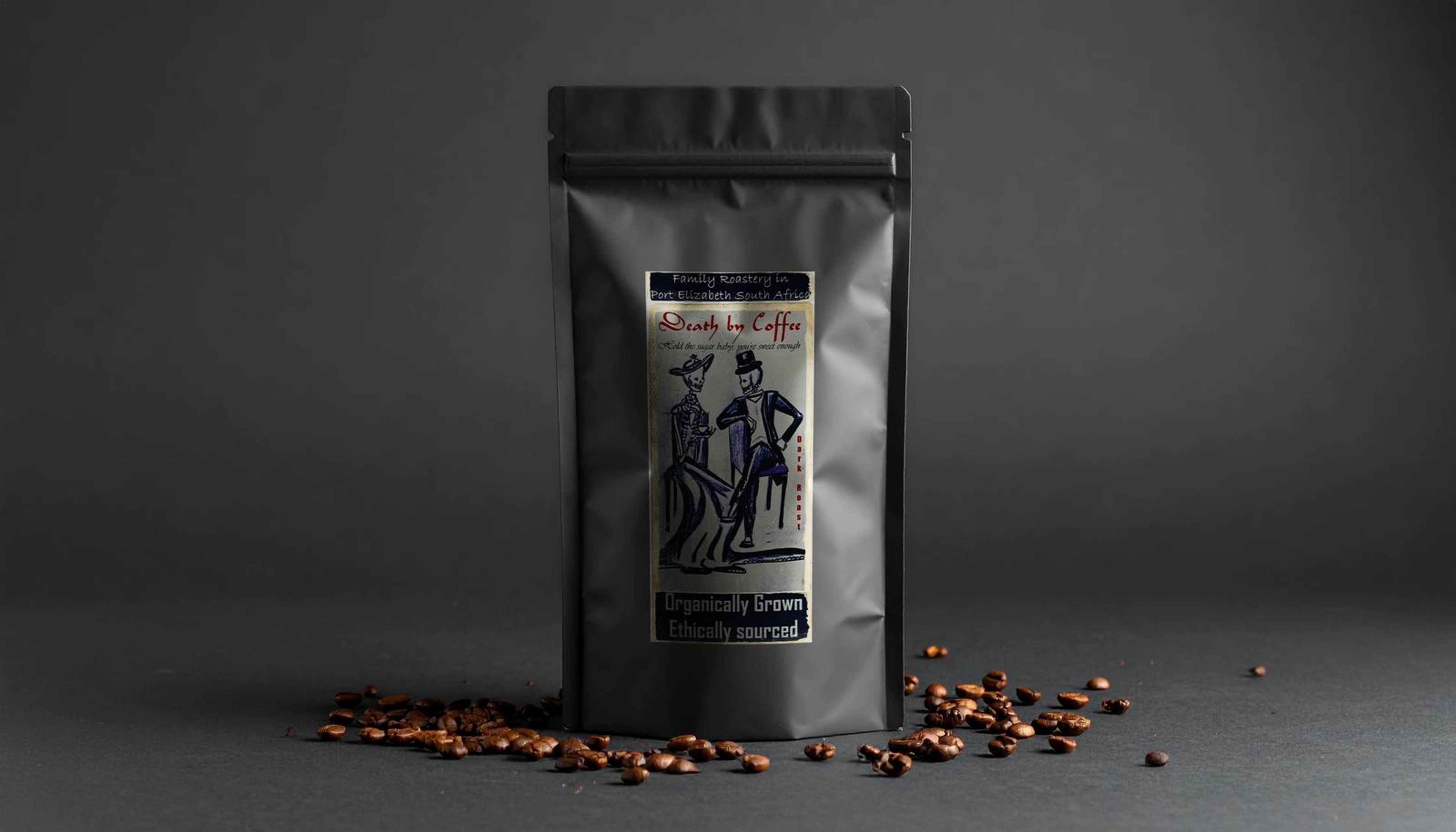 A Death By Coffee coffee bag on a dark studio backdrop with scattered coffee beans