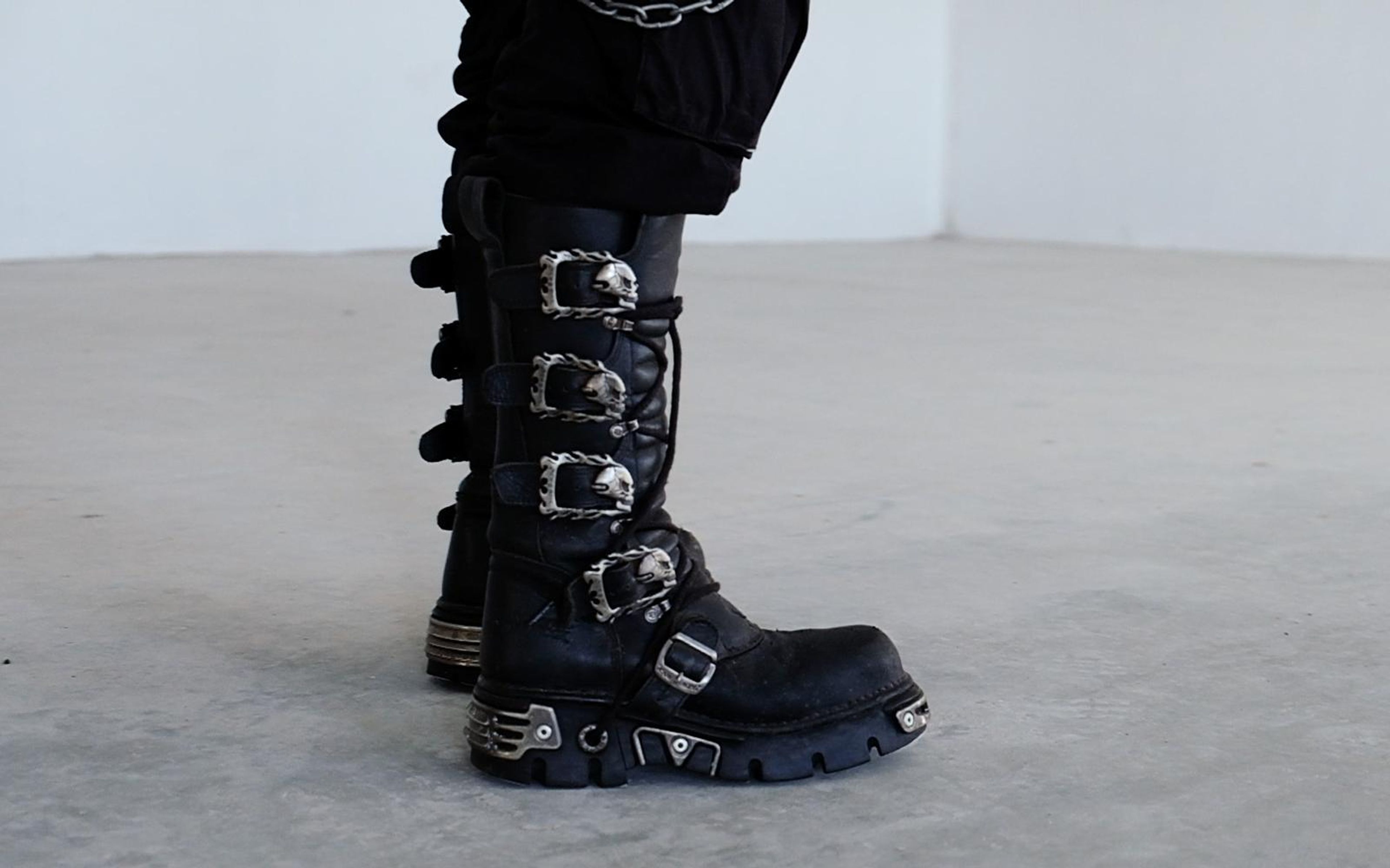 Side view of a tall black boot with four skull buckles and metal heel plate on a grey floor