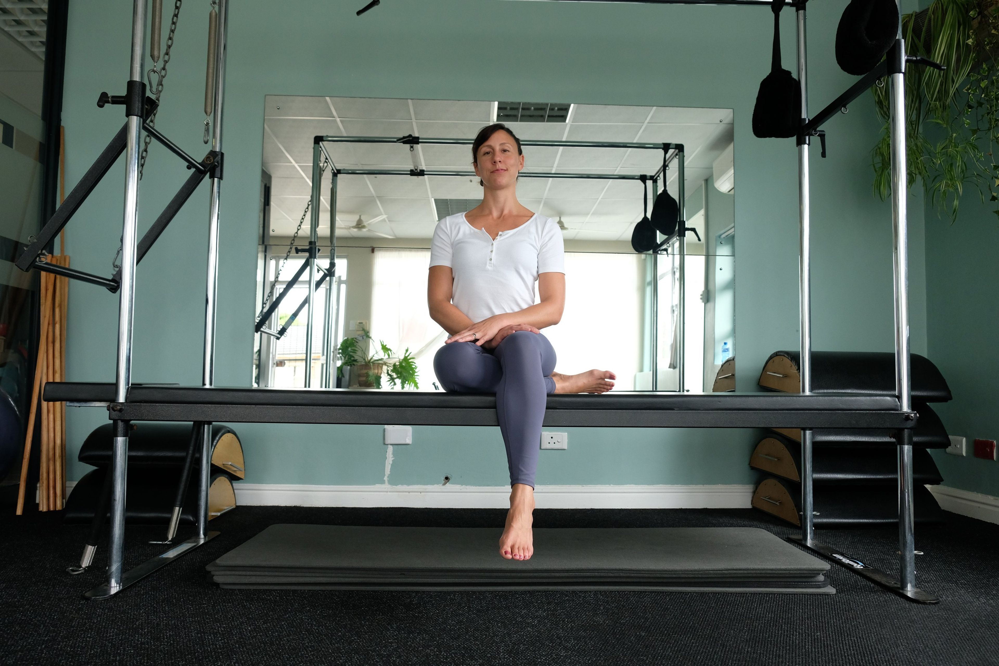 Instructor in a white top and leggings seated on a Pilates reformer bench in the studio