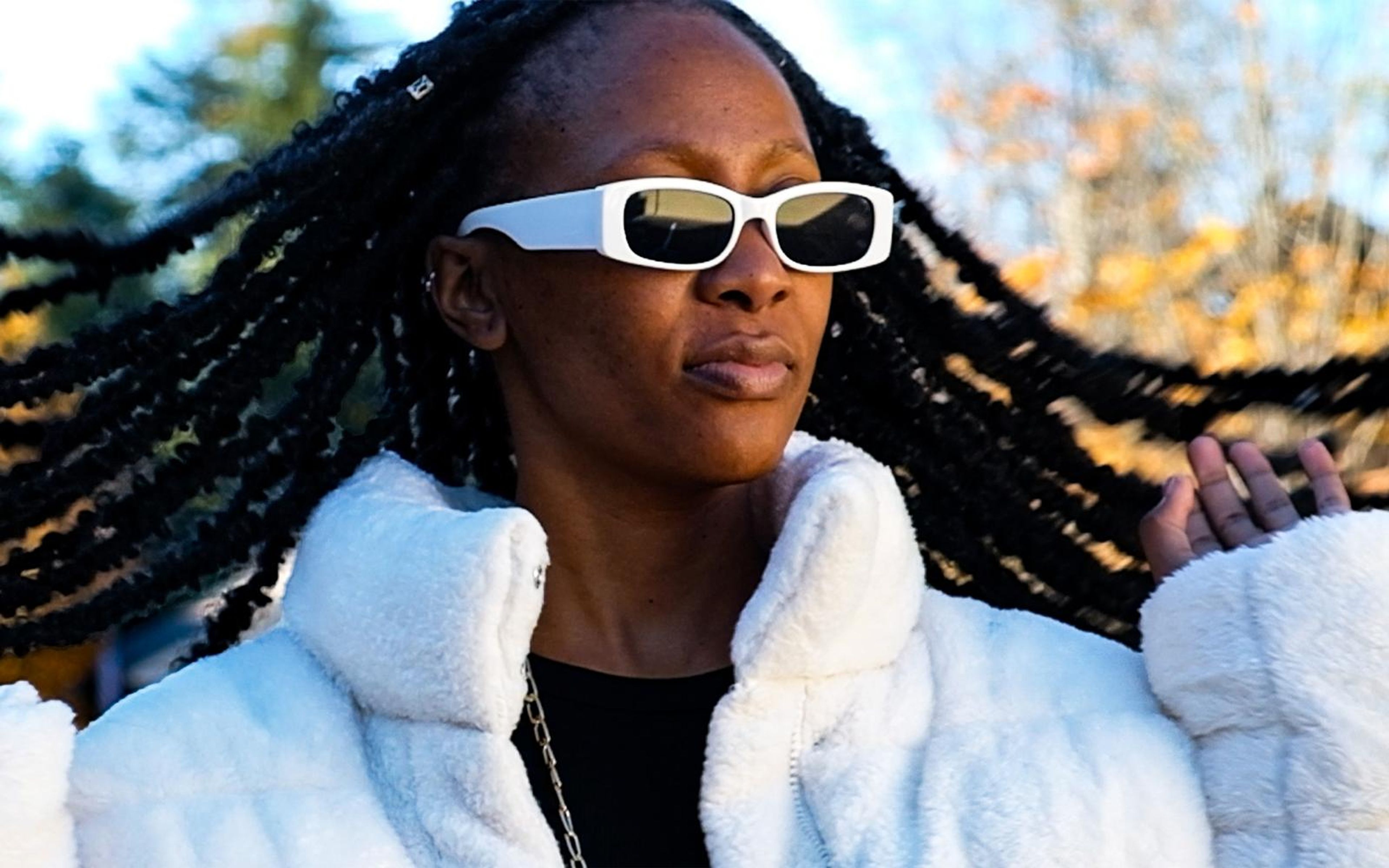 Model flicking her long braids in white sunglasses and a fur jacket, autumn trees behind