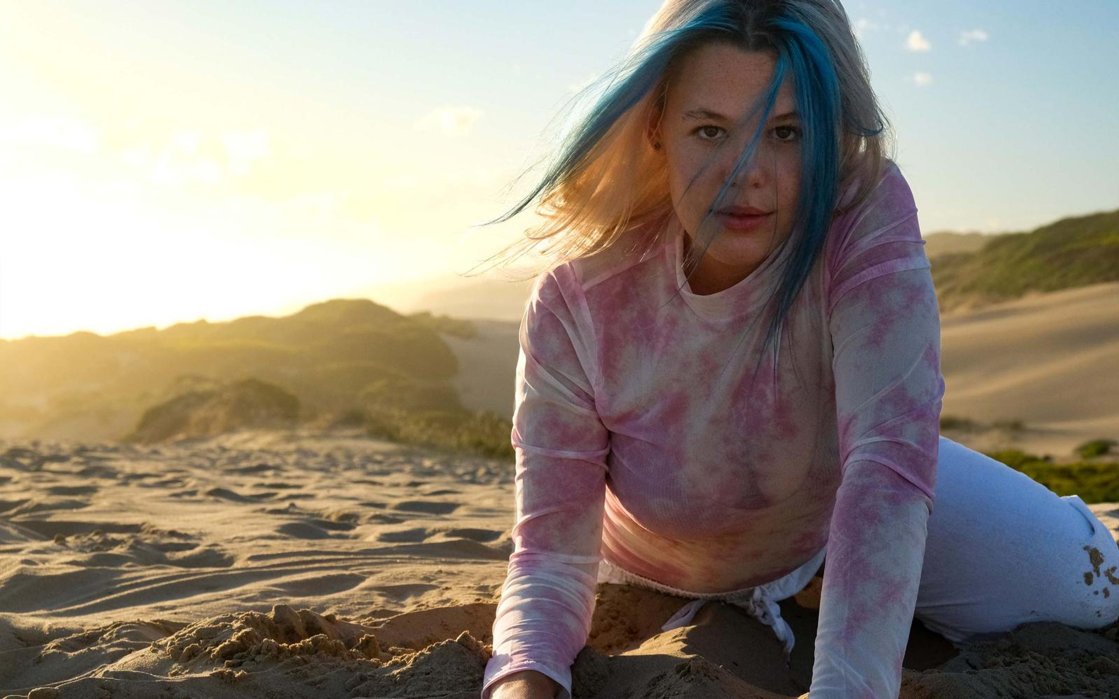 Woman kneeling on the sand with hair lifted by wind, looking into the lens