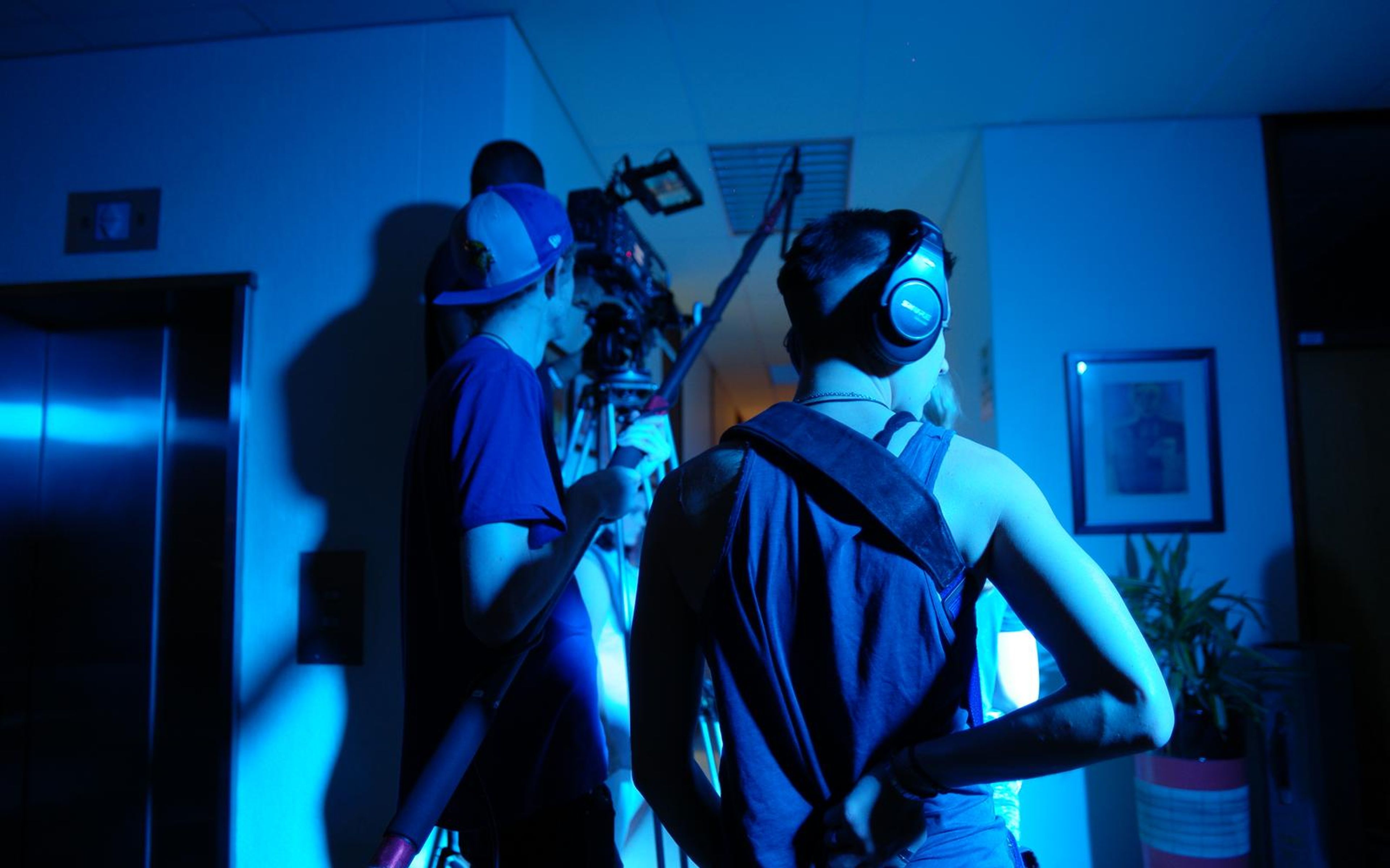 Crew in blue light beside a camera on a tripod in a corridor, one wearing headphones