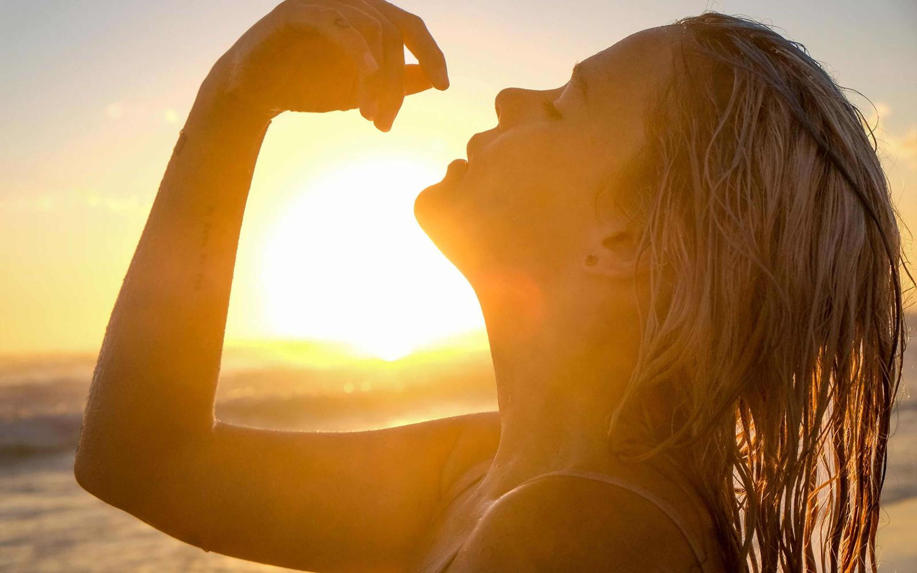 Profile of a woman with wet hair tilting her head back, one arm raised and fist near her face, sun glowing between