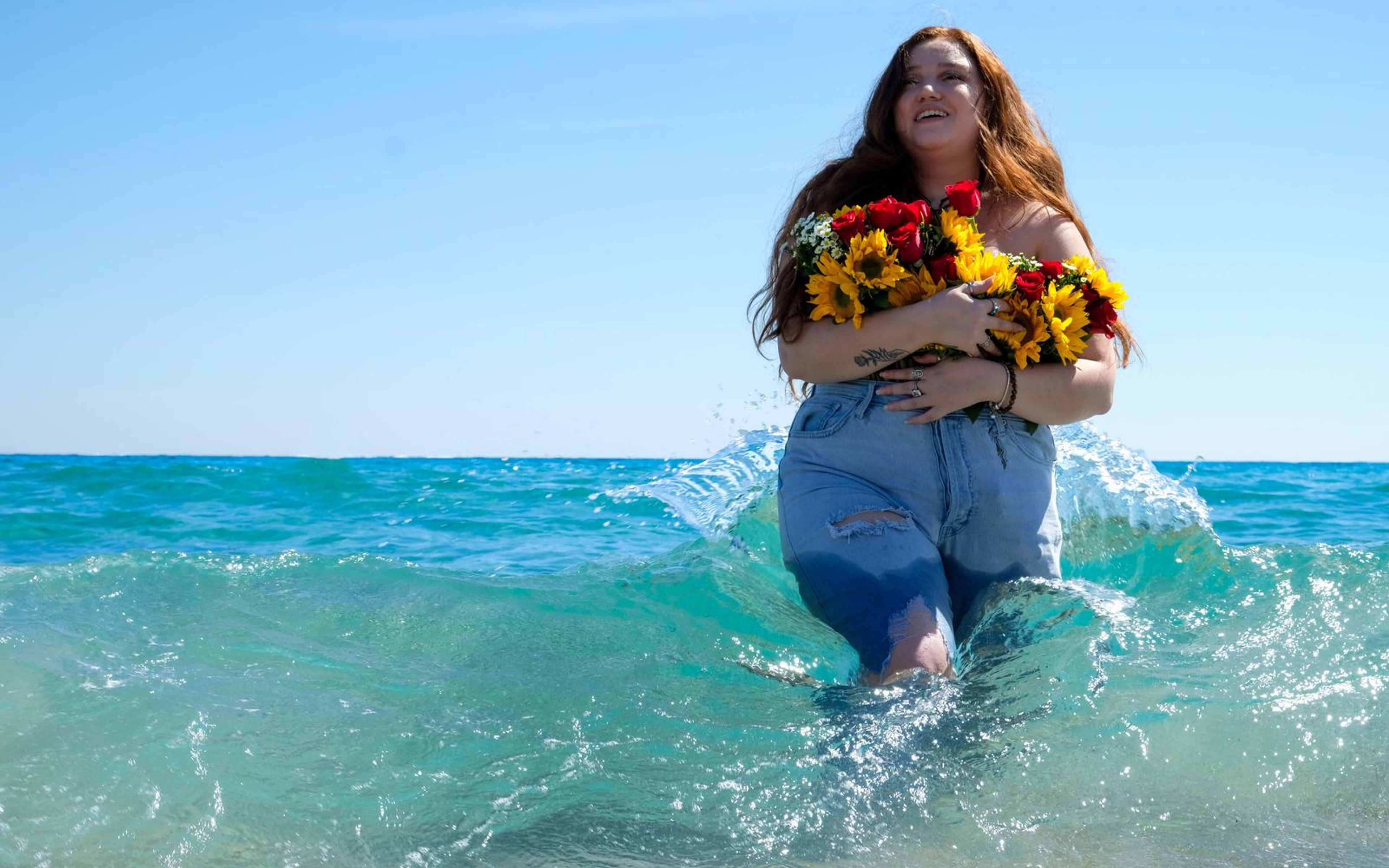 Model with head turned up and a smile, standing in the sea with bouquet, water swirling at her legs