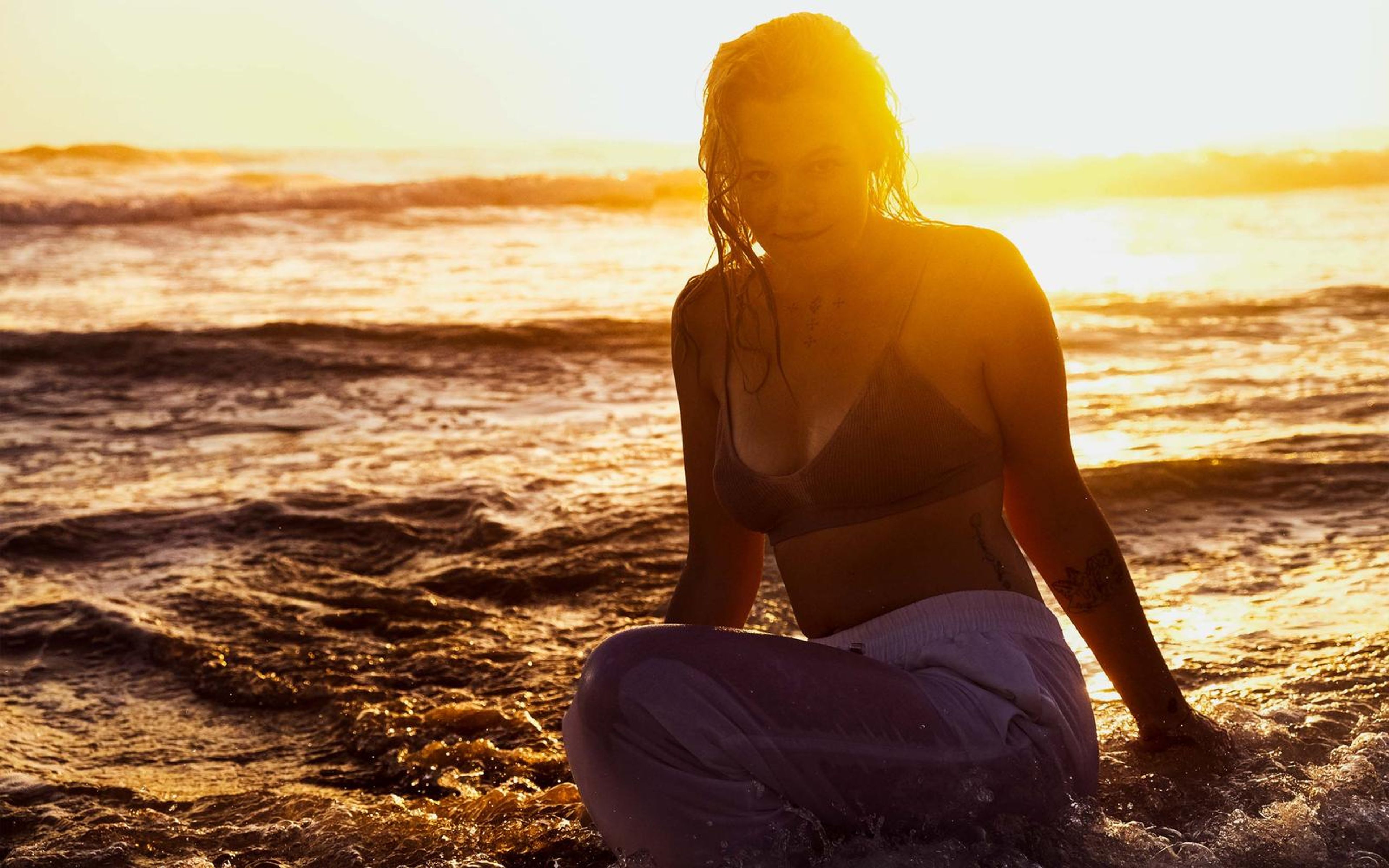 Woman sitting cross-legged in glowing amber water, bikini top, wet hair, looking down, deep orange sunset
