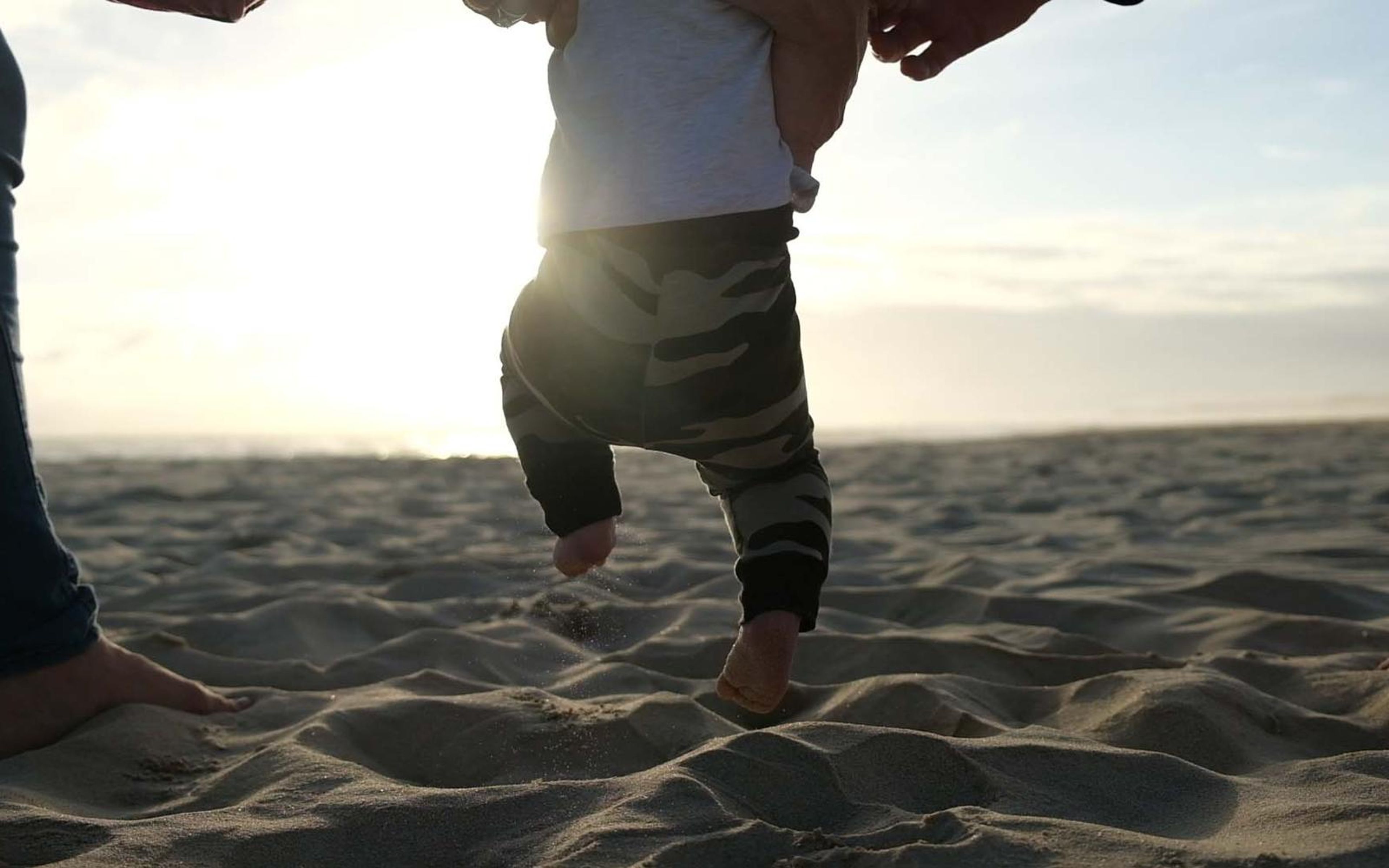 Parent holding a toddler's hands as the child takes steps on a sunlit sandy beach