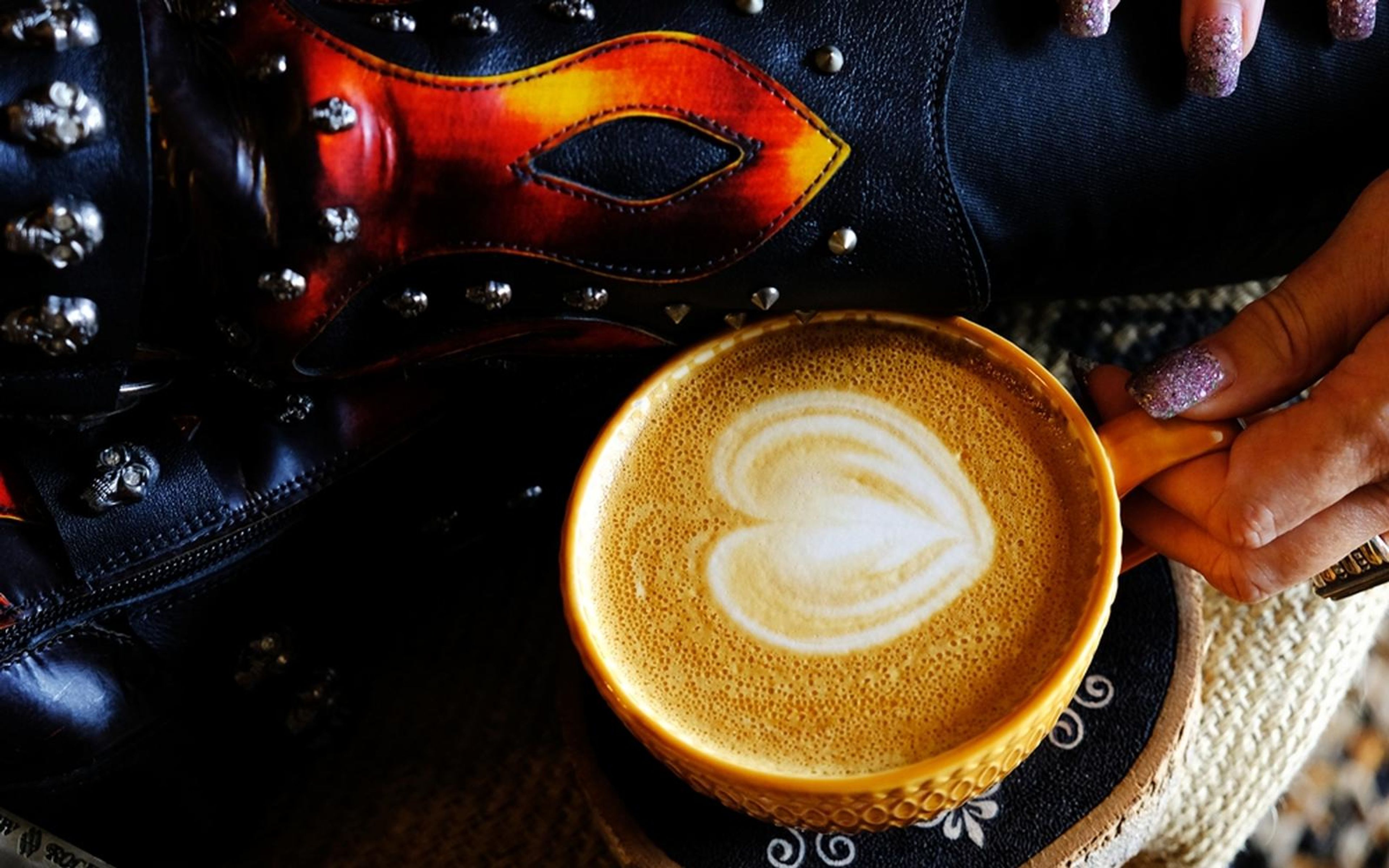 Overhead view of heart-shaped latte art in a yellow cup, studded boot and fingers with painted nails