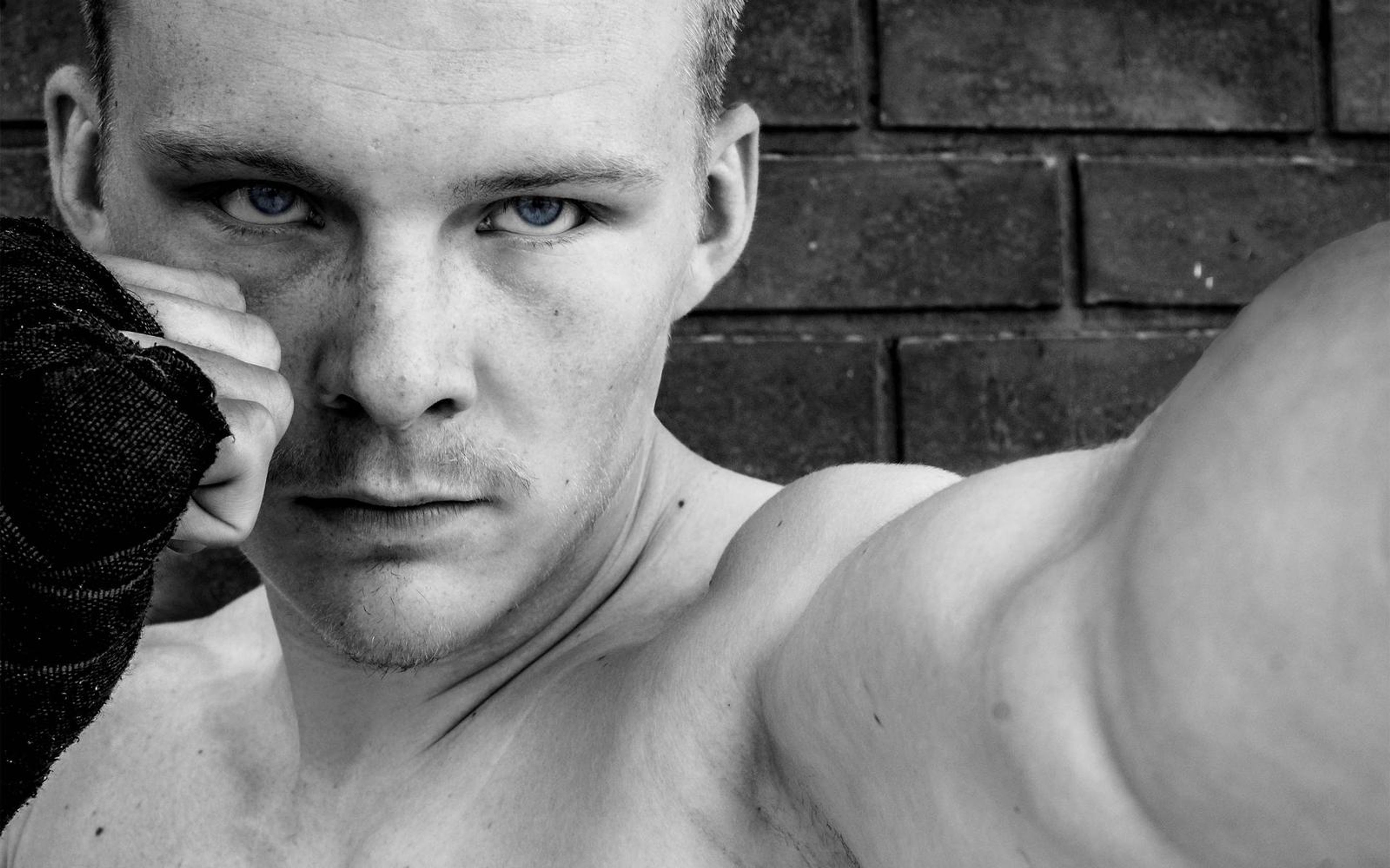 Black and white close portrait of a man with a hard stare, wrapped fist raised beside his face, brick wall behind