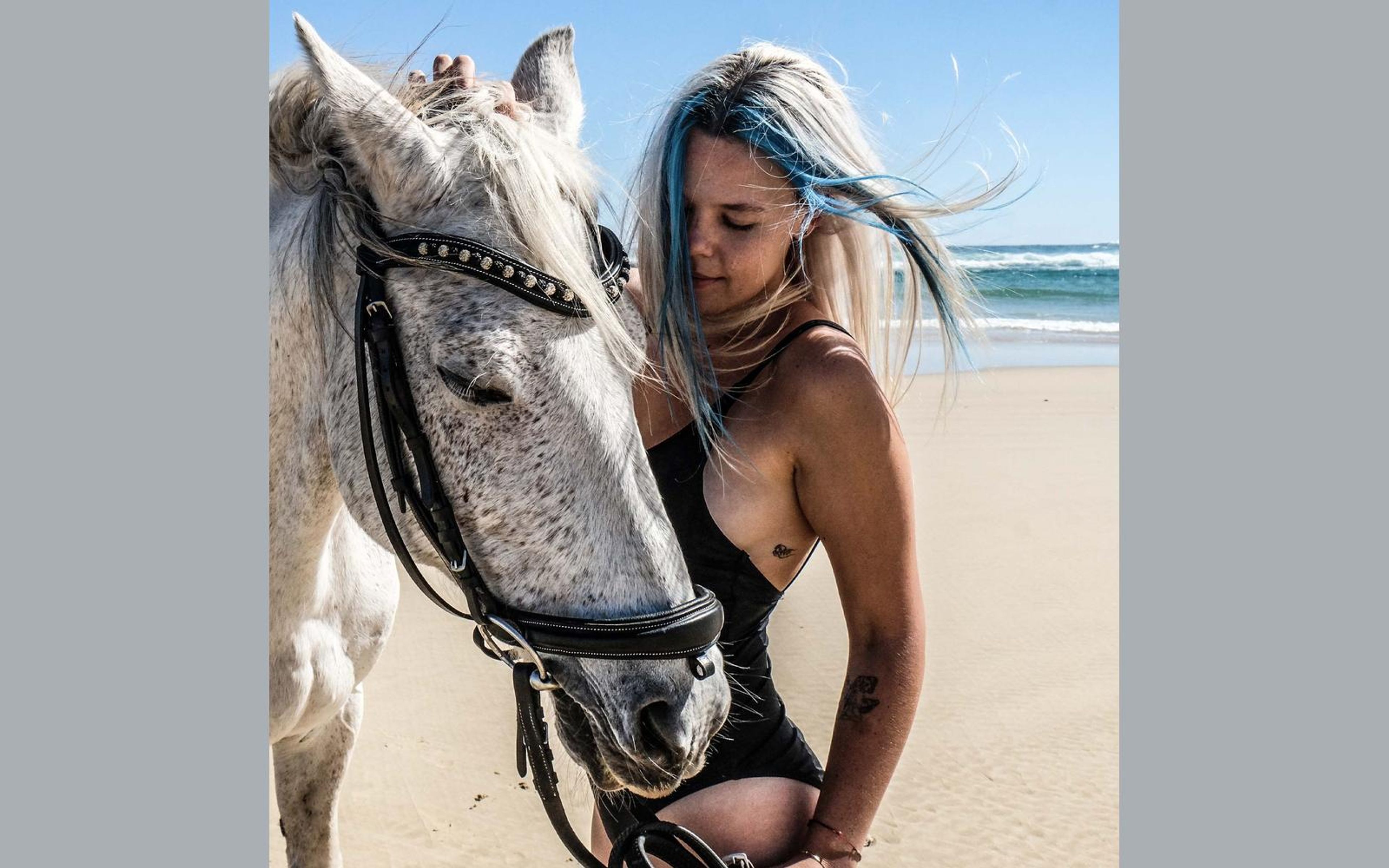 Woman with blue-streaked hair standing beside a white horse and looking down, in a black swimsuit