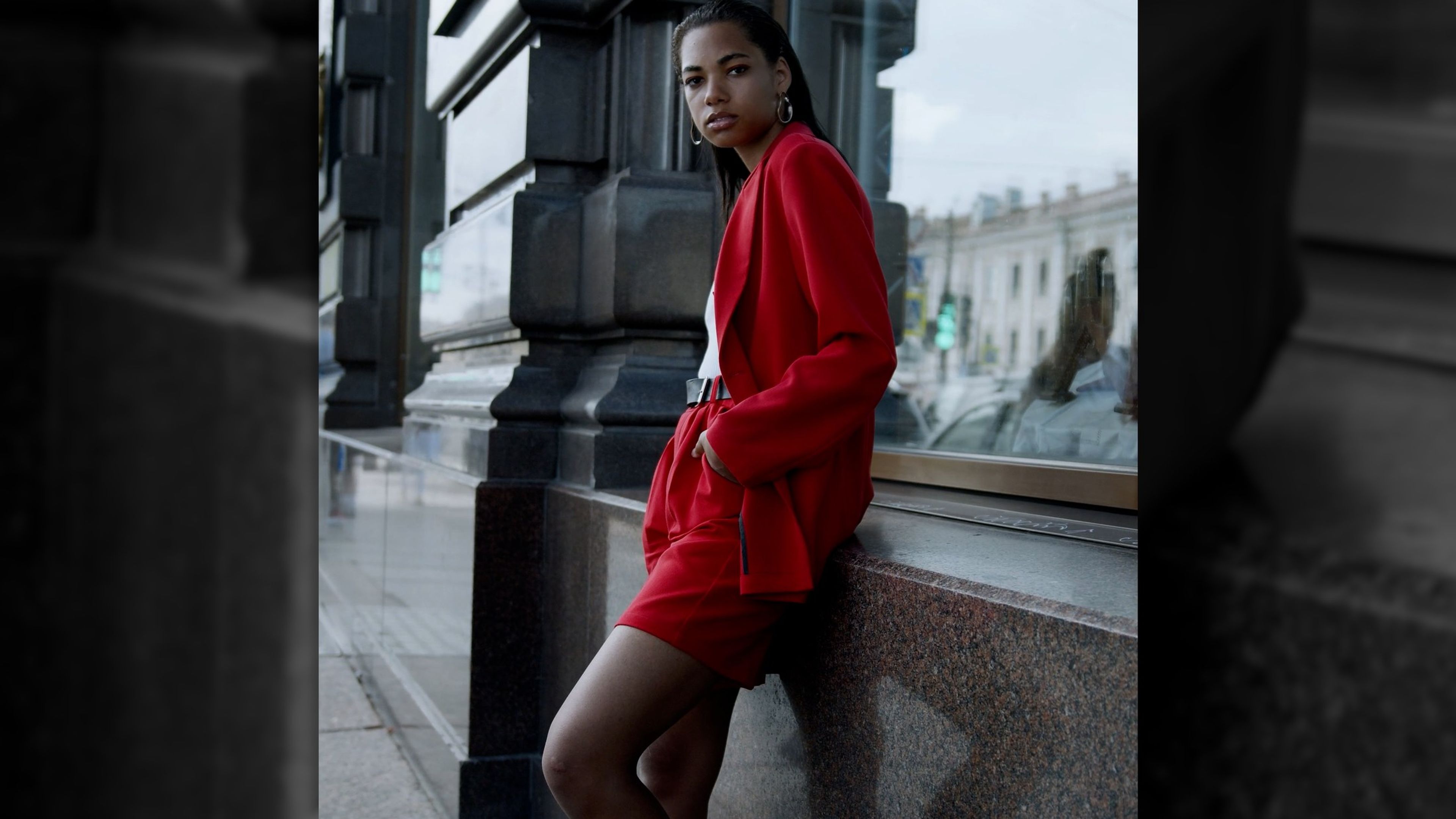 Woman in a red coat leaning against a dark stone wall beside a window