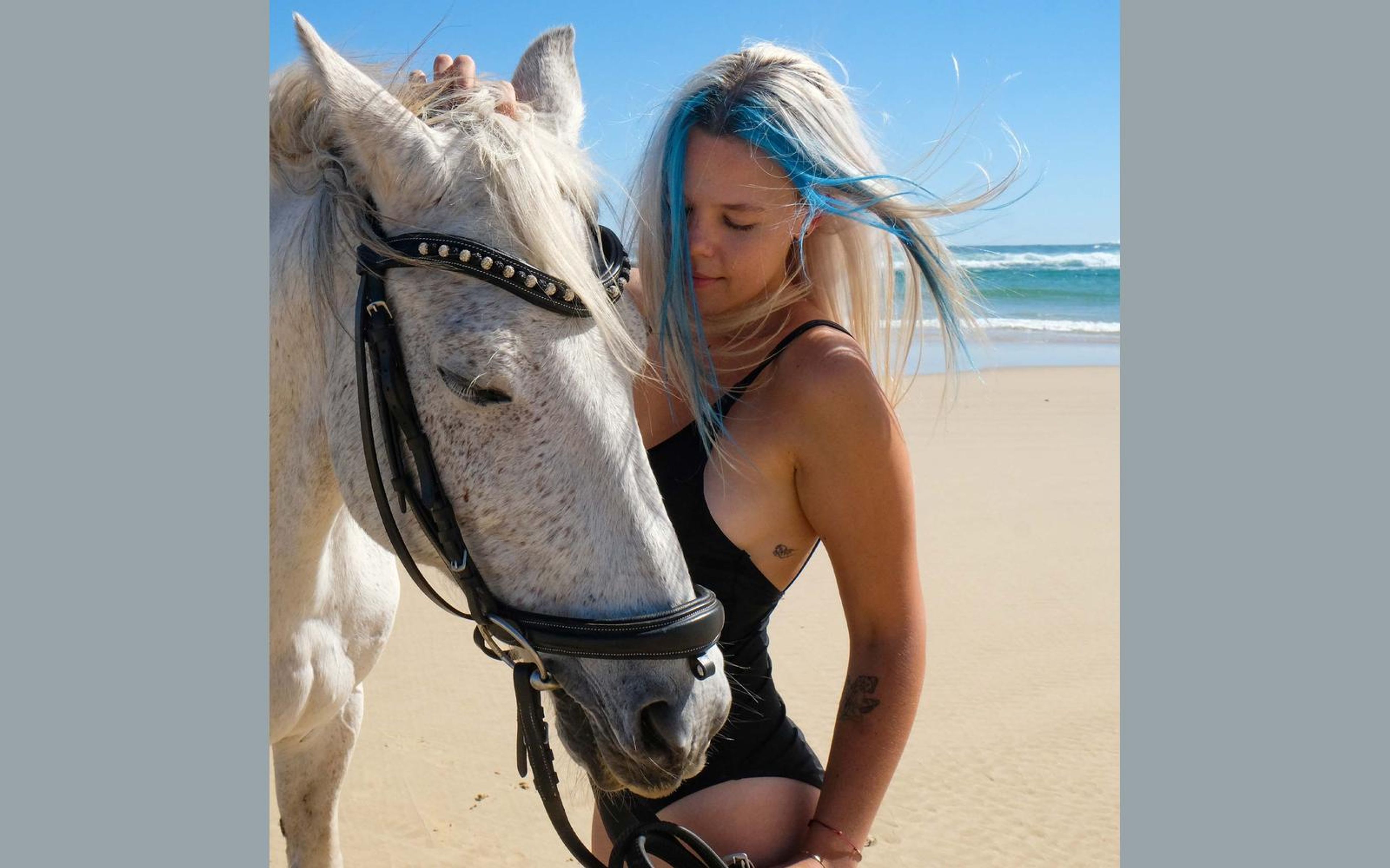 Woman with blue-streaked blonde hair leaning her head against a white horse on a sunny beach, black swimsuit, ocean behind