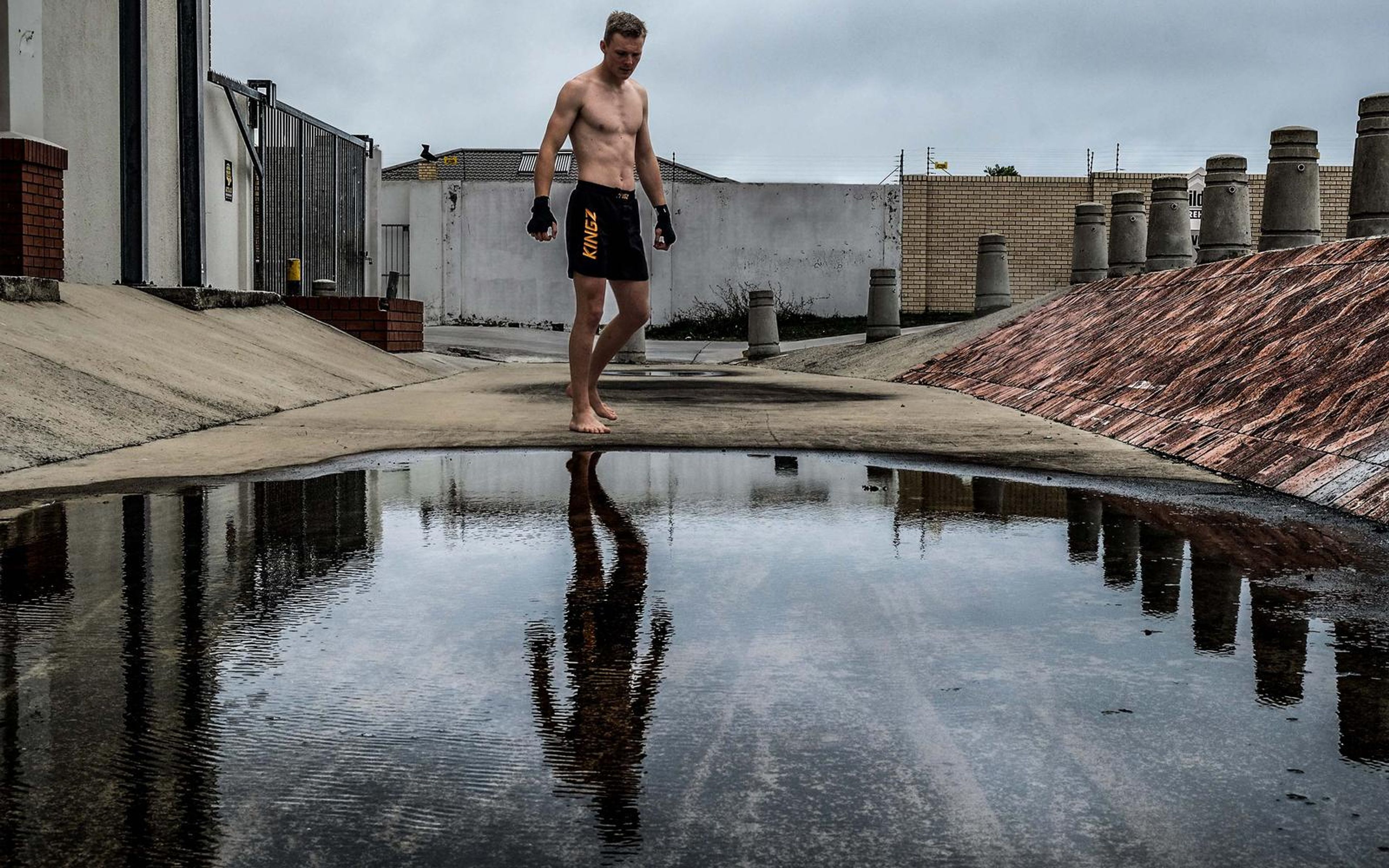 Man standing barefoot at the edge of a puddle, hands wrapped, relaxed stance, overcast sky