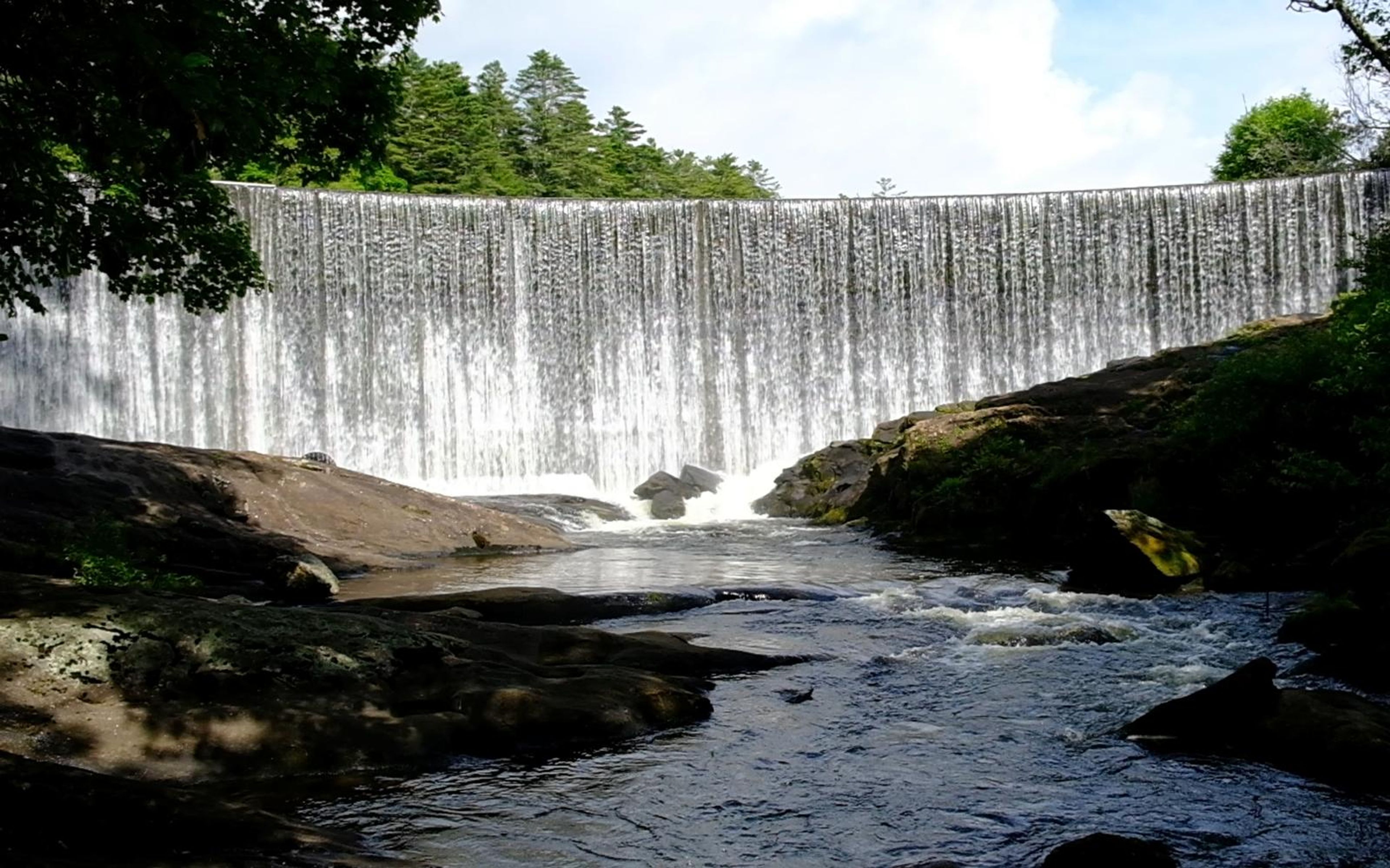Wide view of a long curved waterfall across a river, with sunlit rocks and dark trees in the foreground