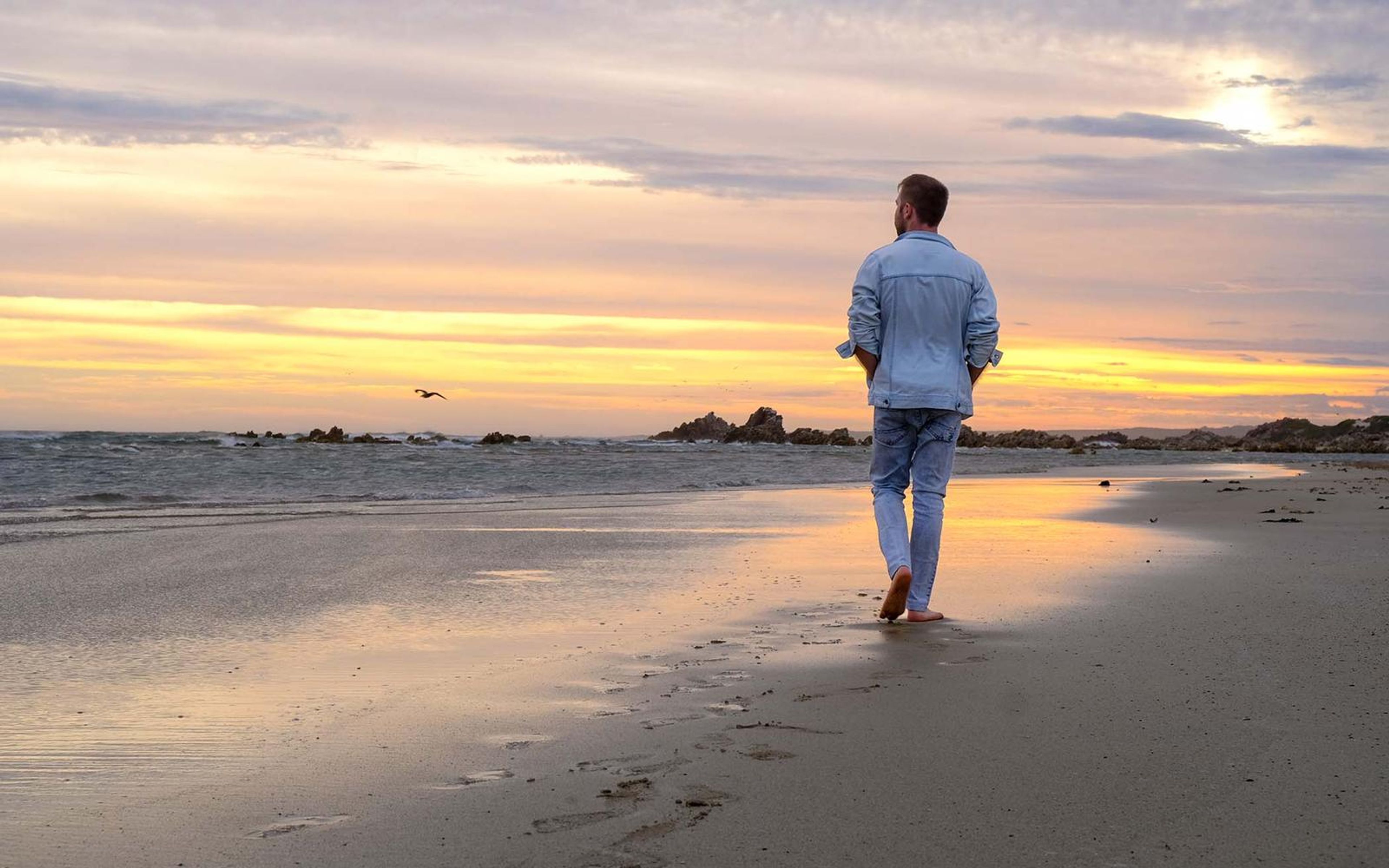 Man walking barefoot along a beach at sunset, reflections in the wet sand, denim jacket