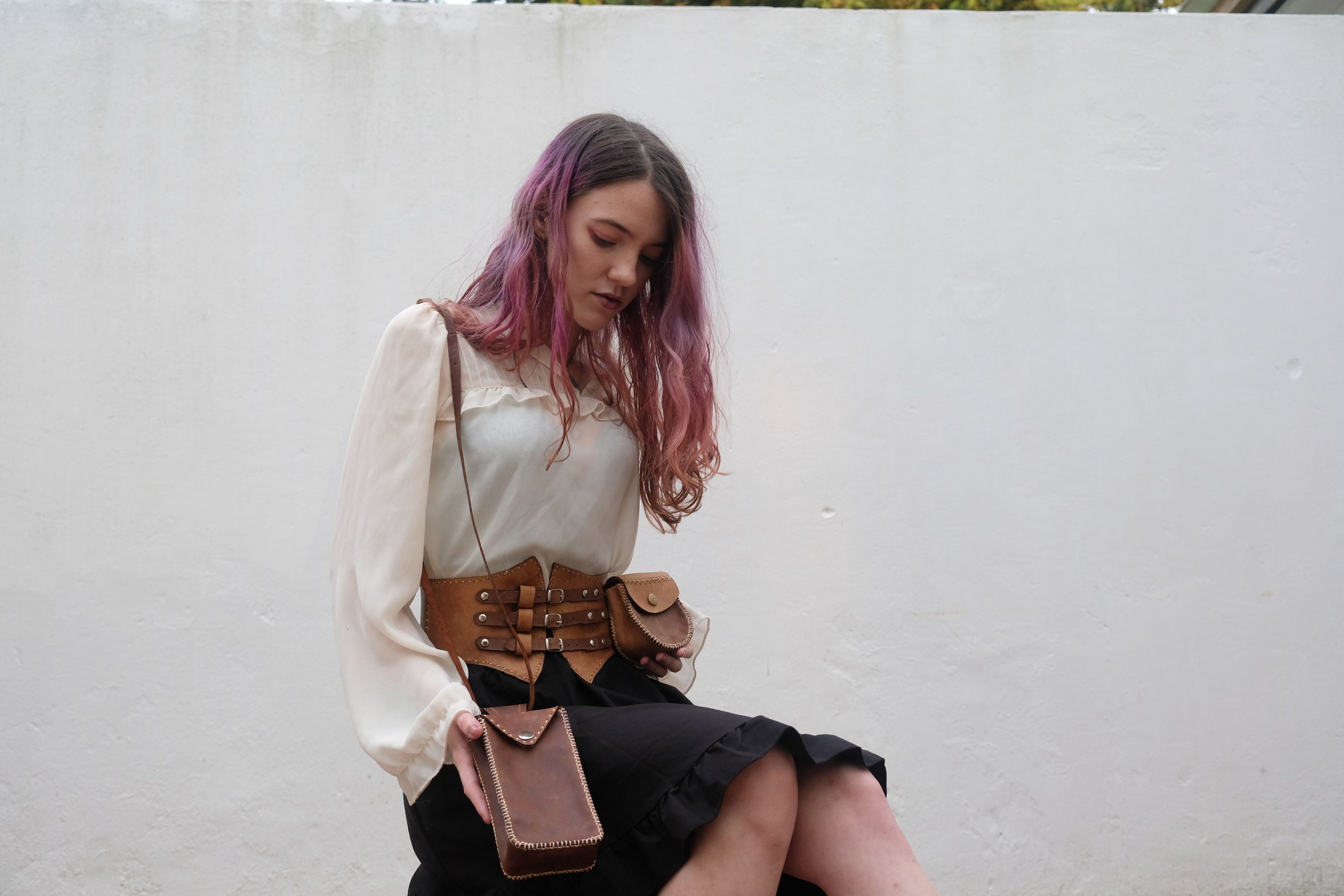 Seated woman with pink hair, wearing a leather corset belt and holding a phone bag