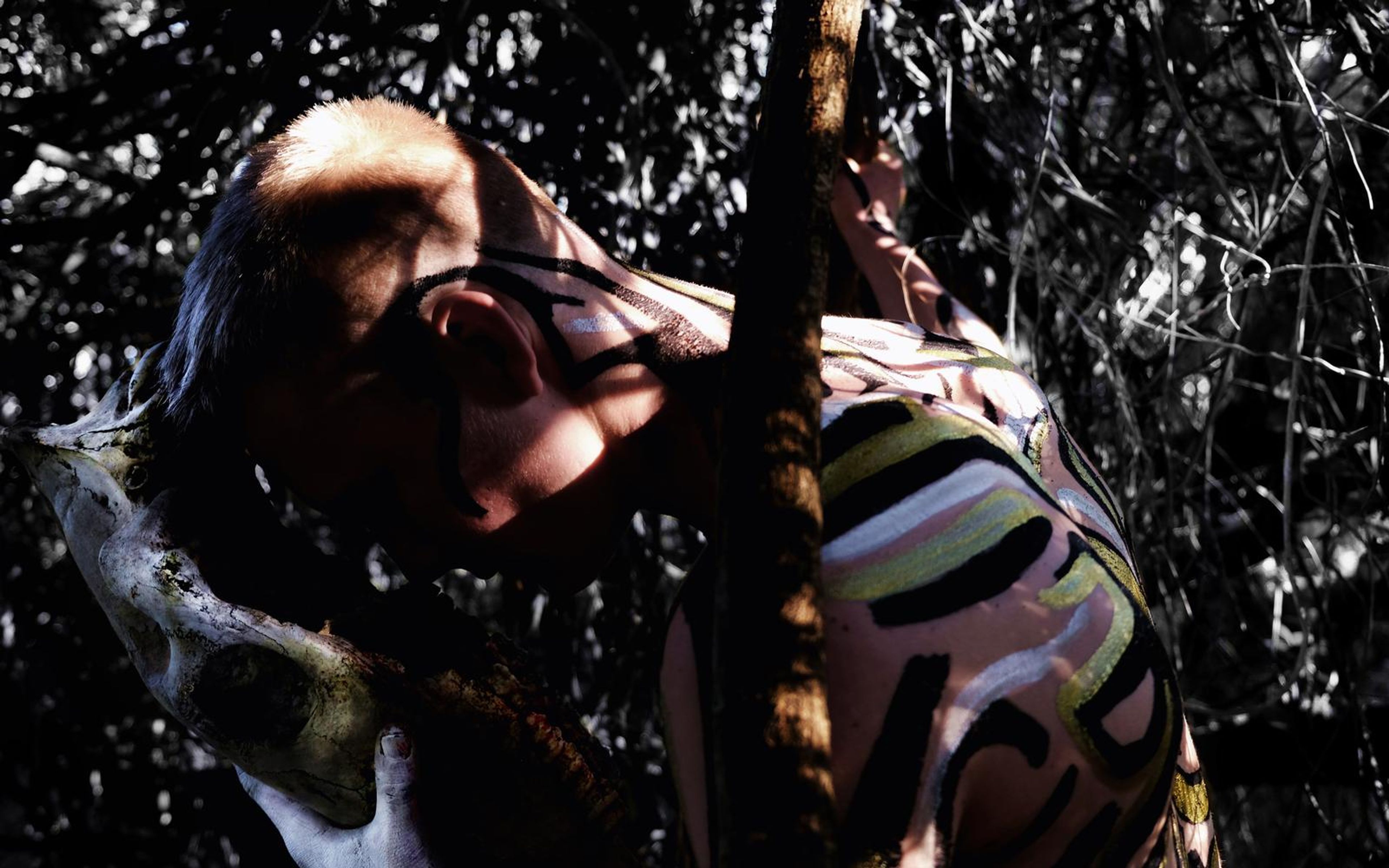 Painted performer hanging head against a thicket of bare branches with a bleached skull held beside the face