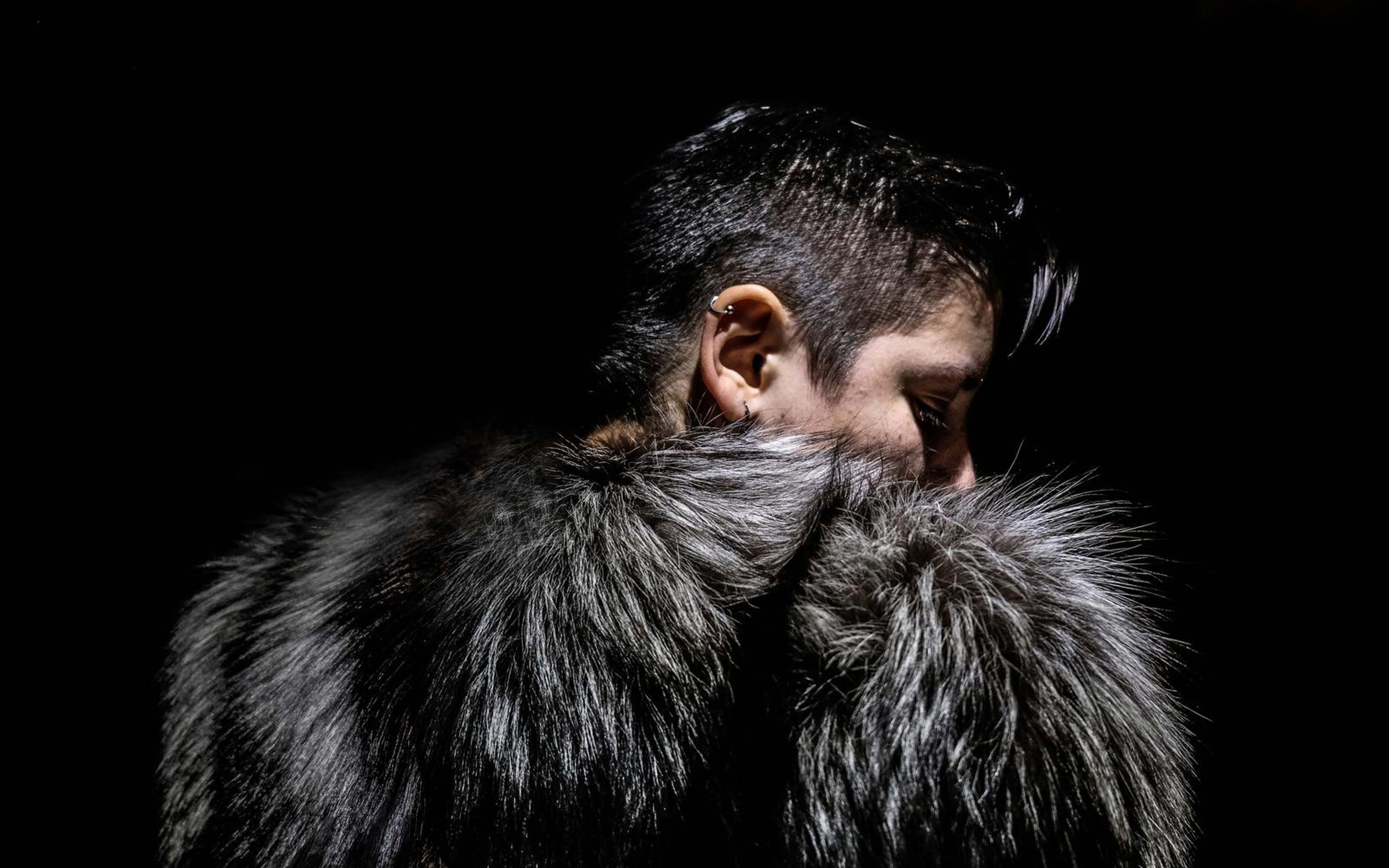 Profile of a person with short dark hair, face turned down into a silver-grey fur collar, lit from the side against black