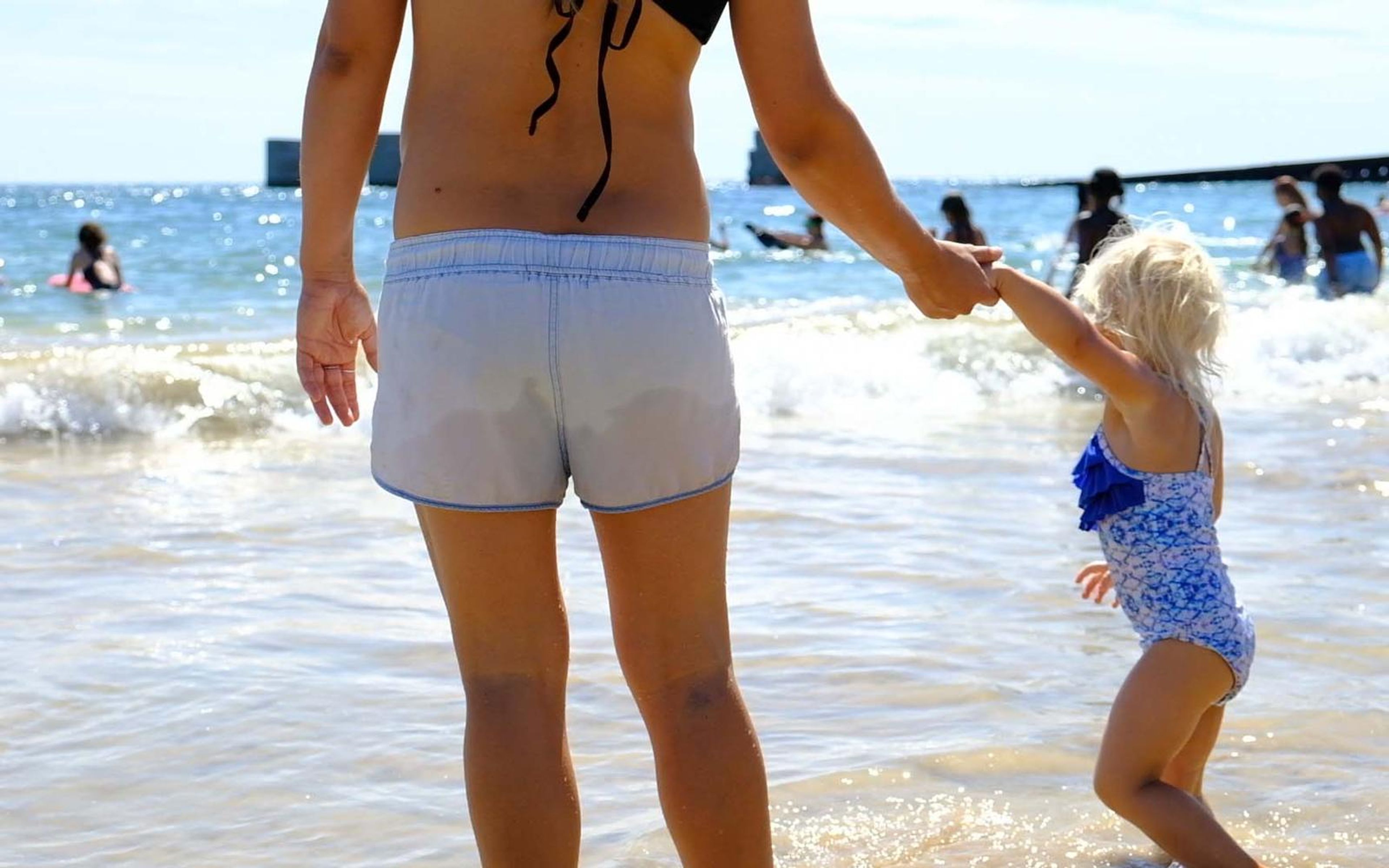 Woman in light shorts holding a small girl's hand as the girl in a blue swimsuit steps into shallow surf
