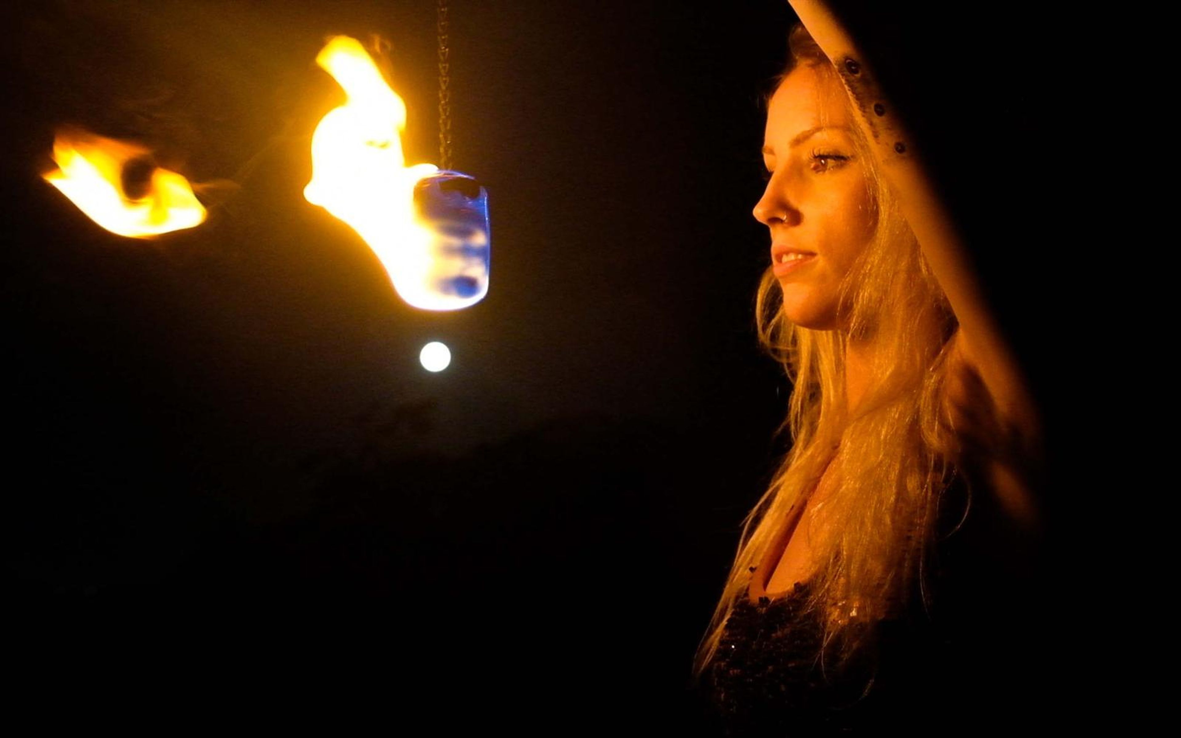Woman in profile at night with a fire poi glowing beside her and a bright light behind, warm side light on her hair