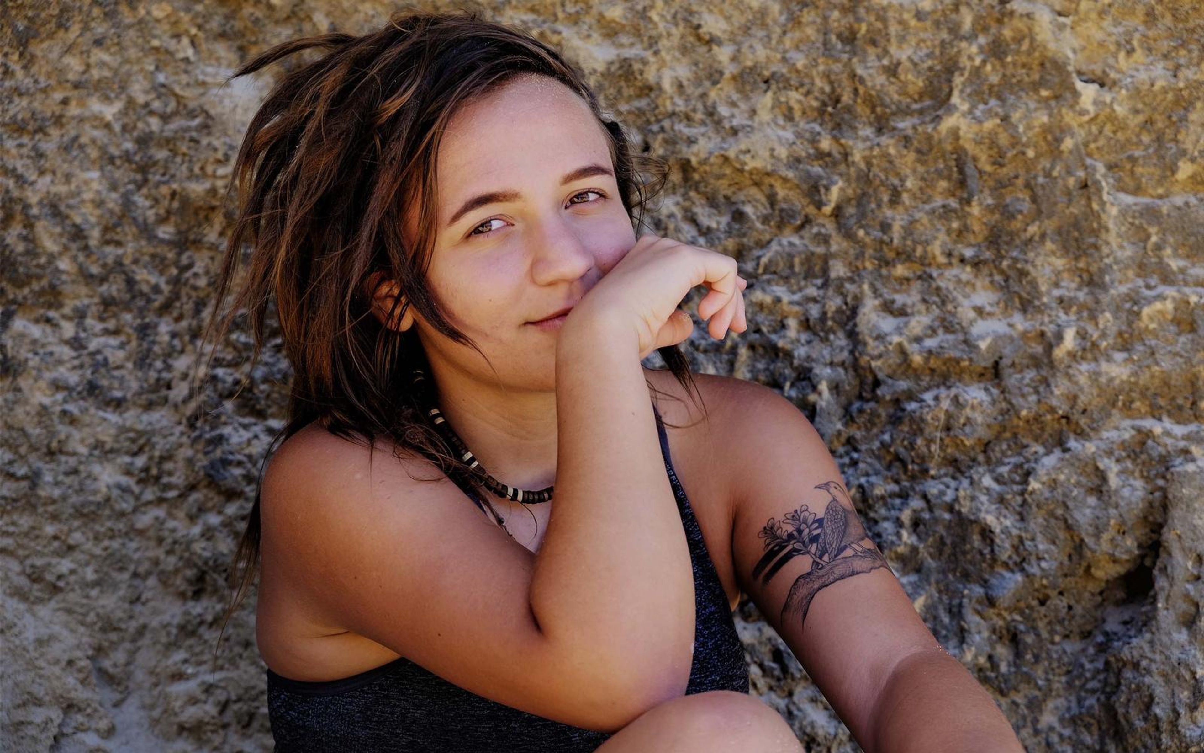 Smiling woman with dreadlocks and a tattooed arm, chin on her hand, seated against a sandy rock wall