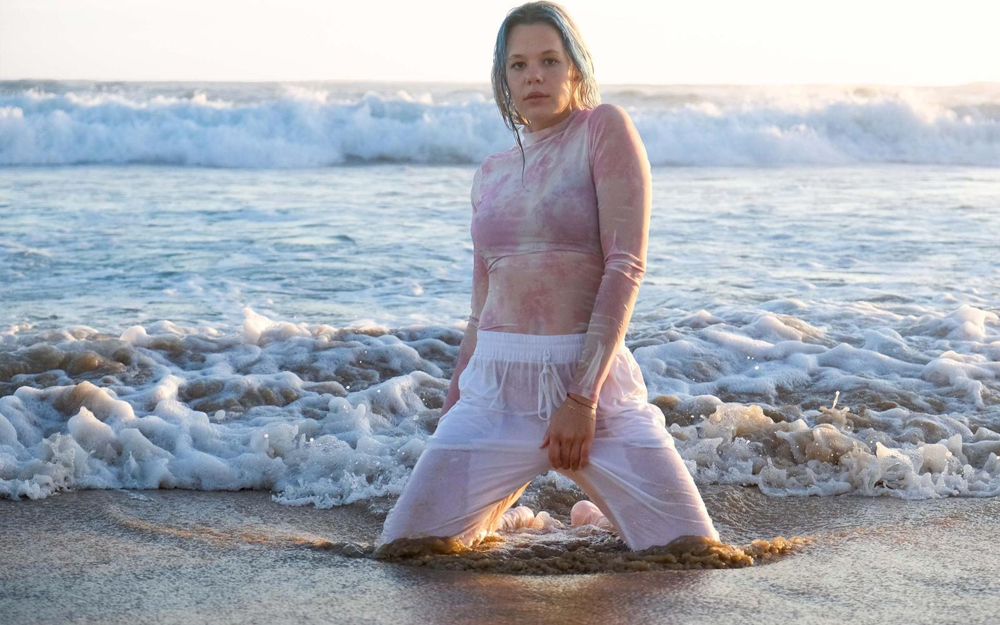 Woman kneeling in churning foam with hands on her thighs, wet pink top, looking straight at the camera