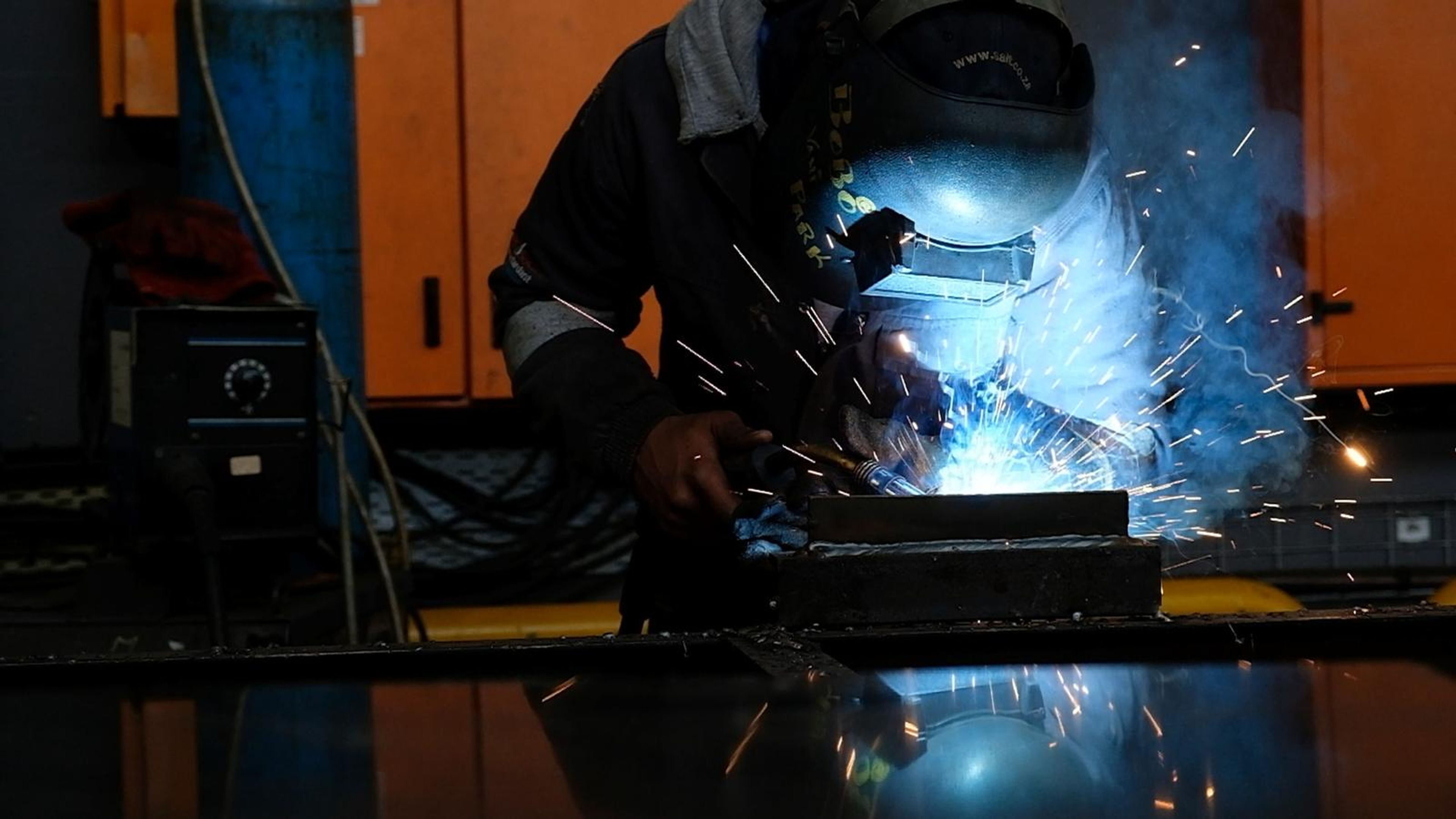 Welder at a steel bar in blue light with sparks drifting past orange cabinets