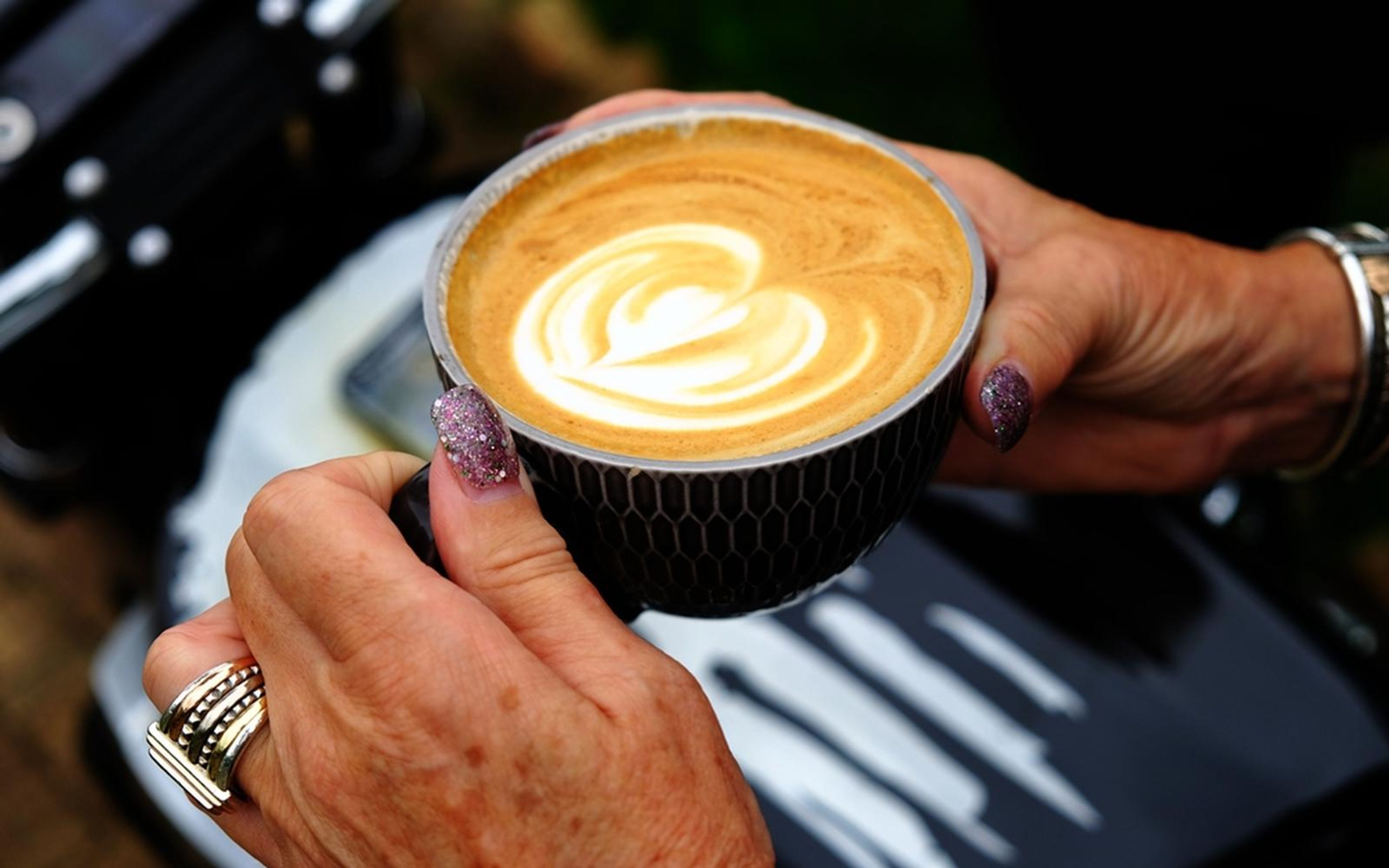 Hands holding a cup of latte with a heart in the foam, ring and glittery nails