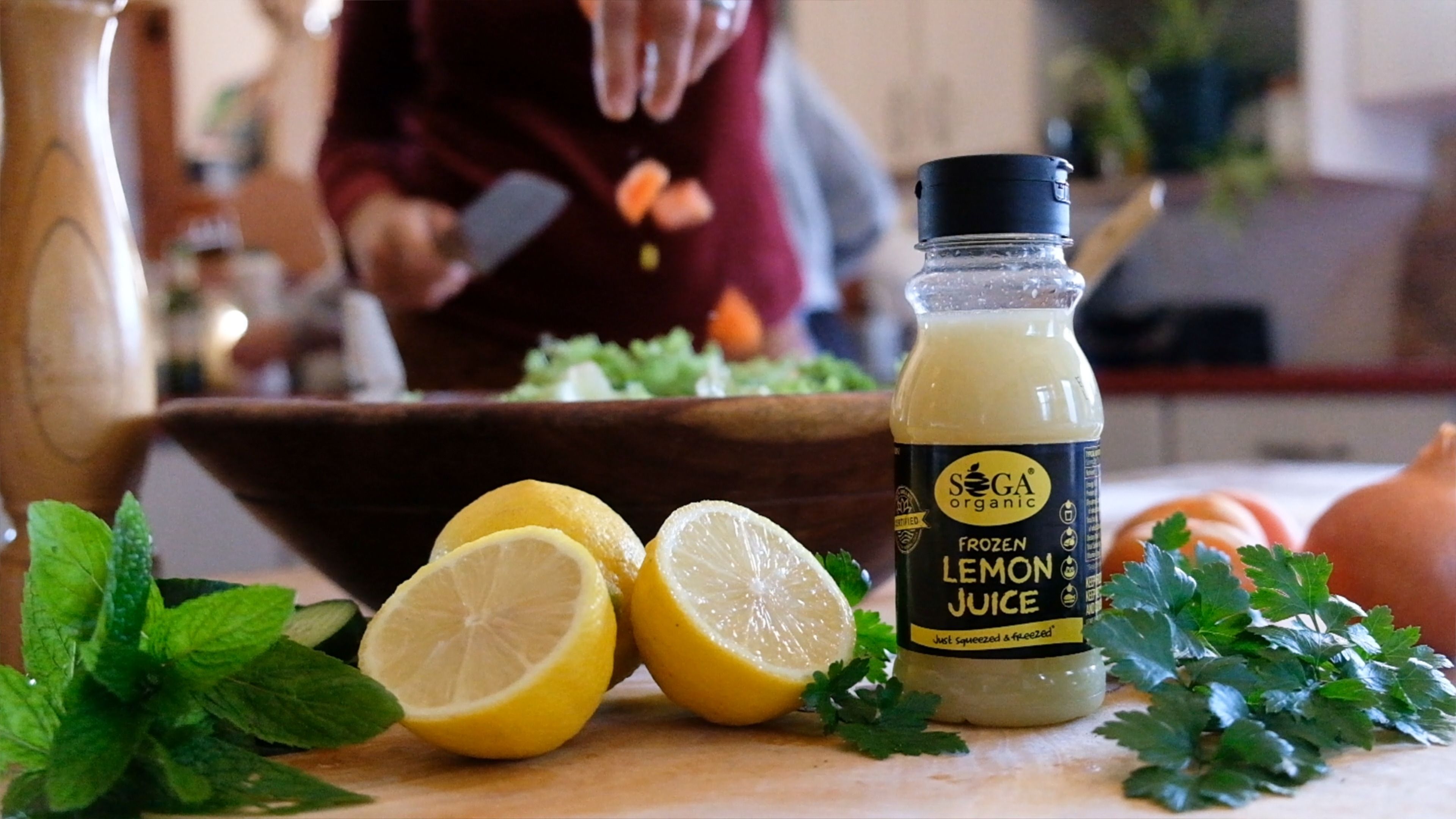 SOGA frozen lemon juice bottle on a kitchen counter with halved lemons and herbs while a salad is prepared