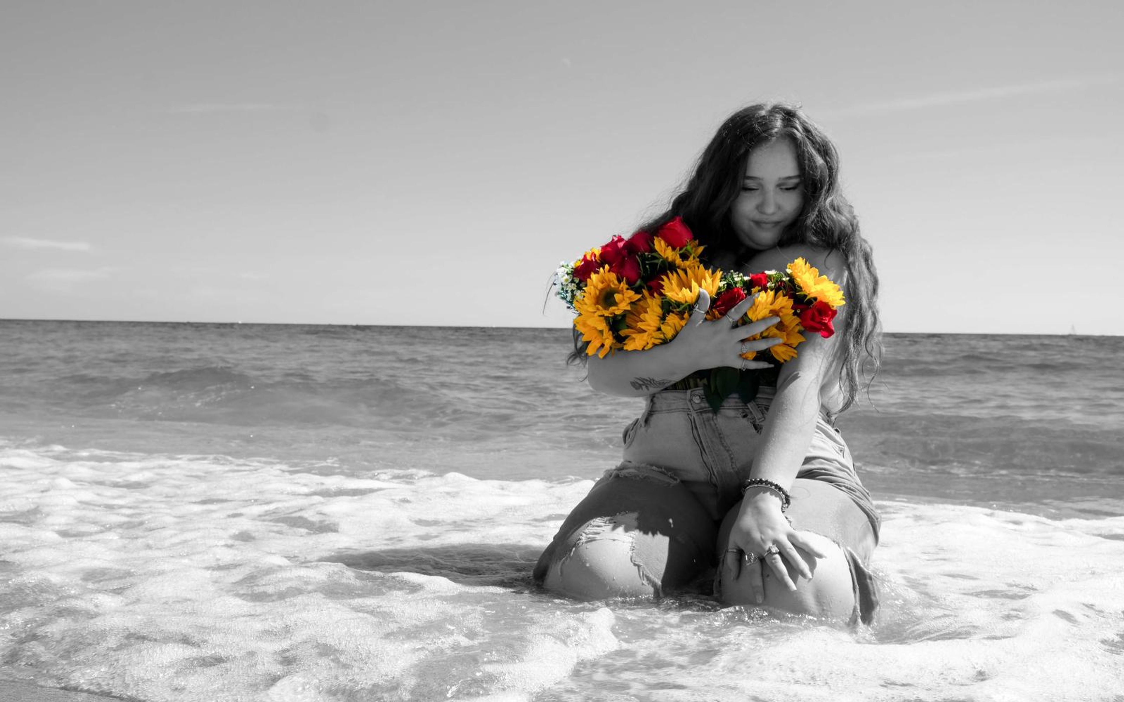 Black and white wide frame of model kneeling in the surf, only the bouquet kept in colour