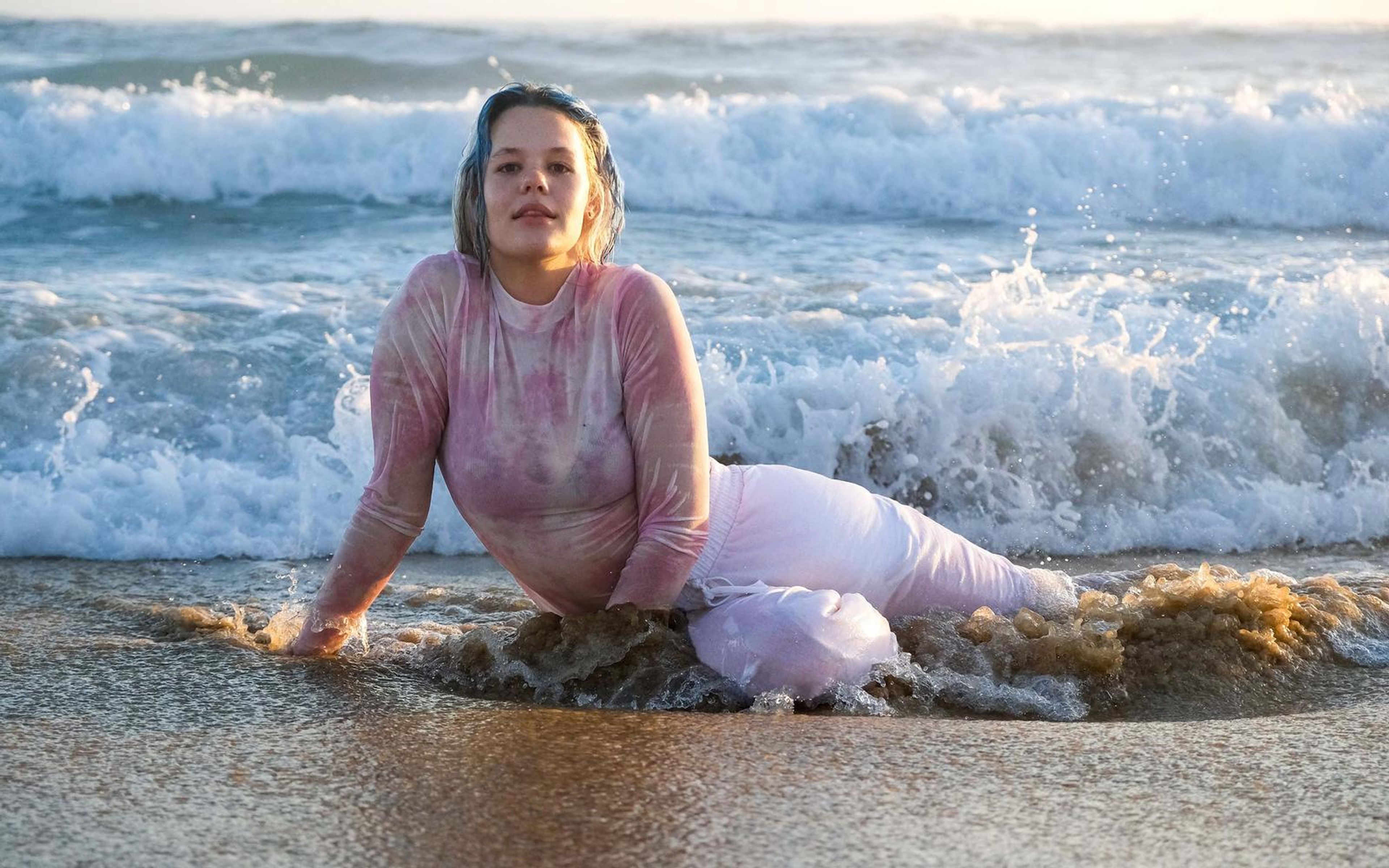 Woman half reclining in foamy surf, hand in the water, looking into the lens, waves breaking behind