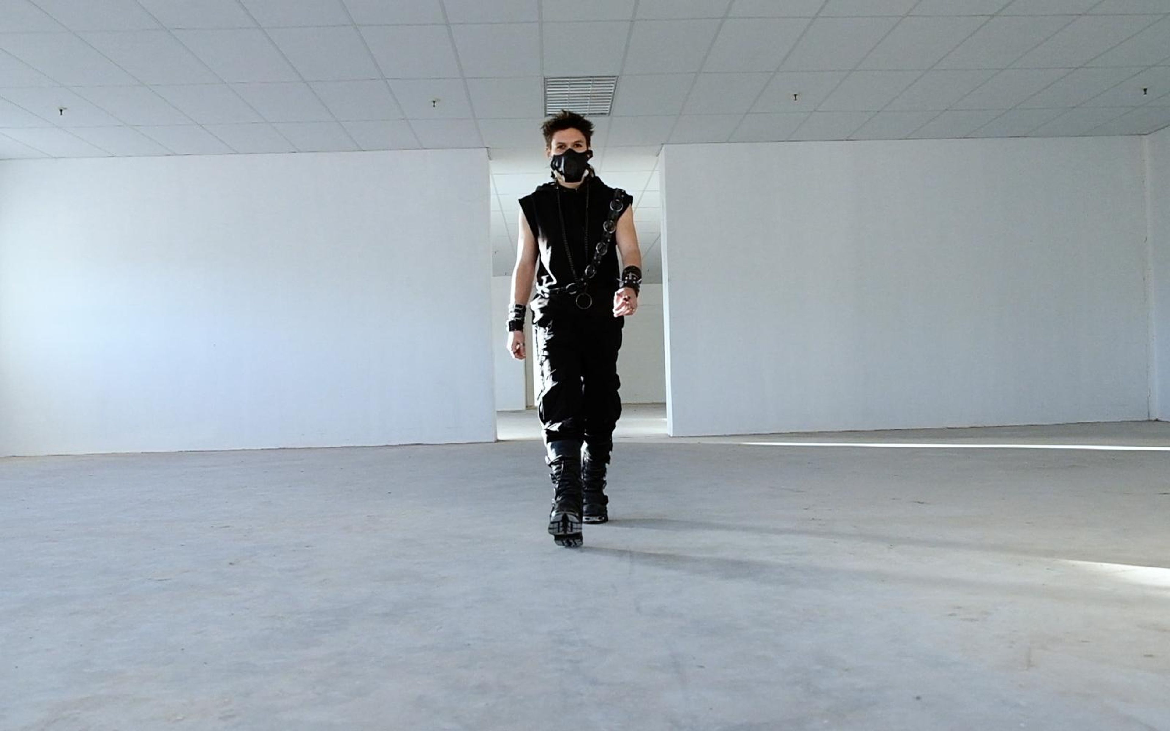 Distant front view of the masked figure in black walking forward in a cool blue-grey empty room