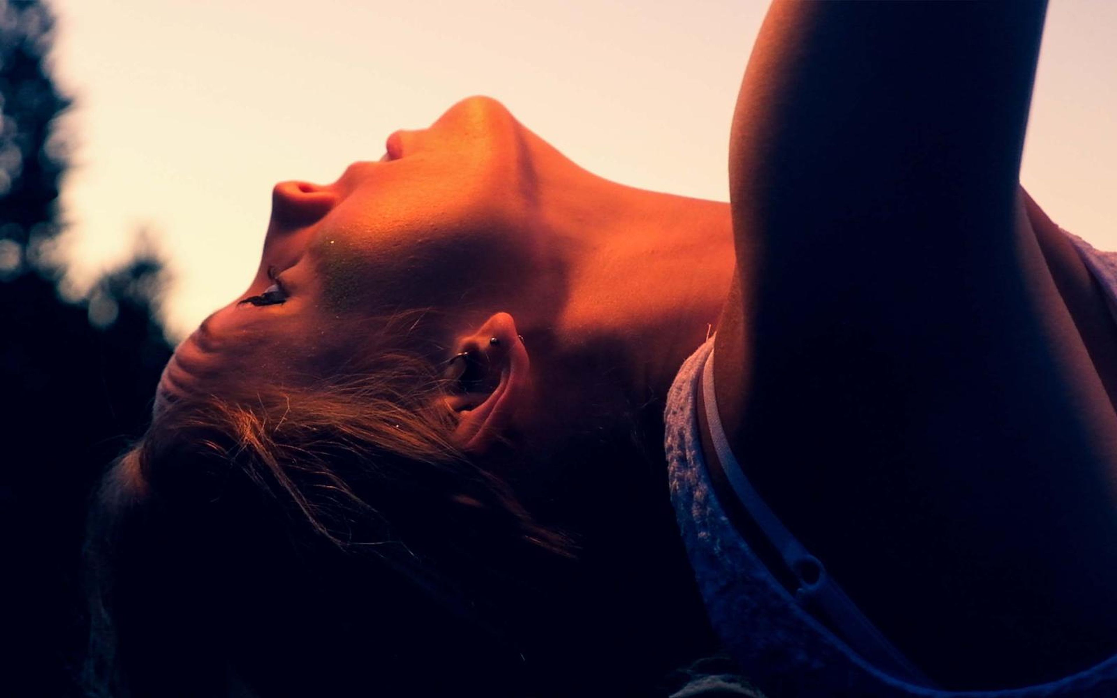 Tighter crop of a woman leaning her head back with an arm raised over her, tree silhouettes behind