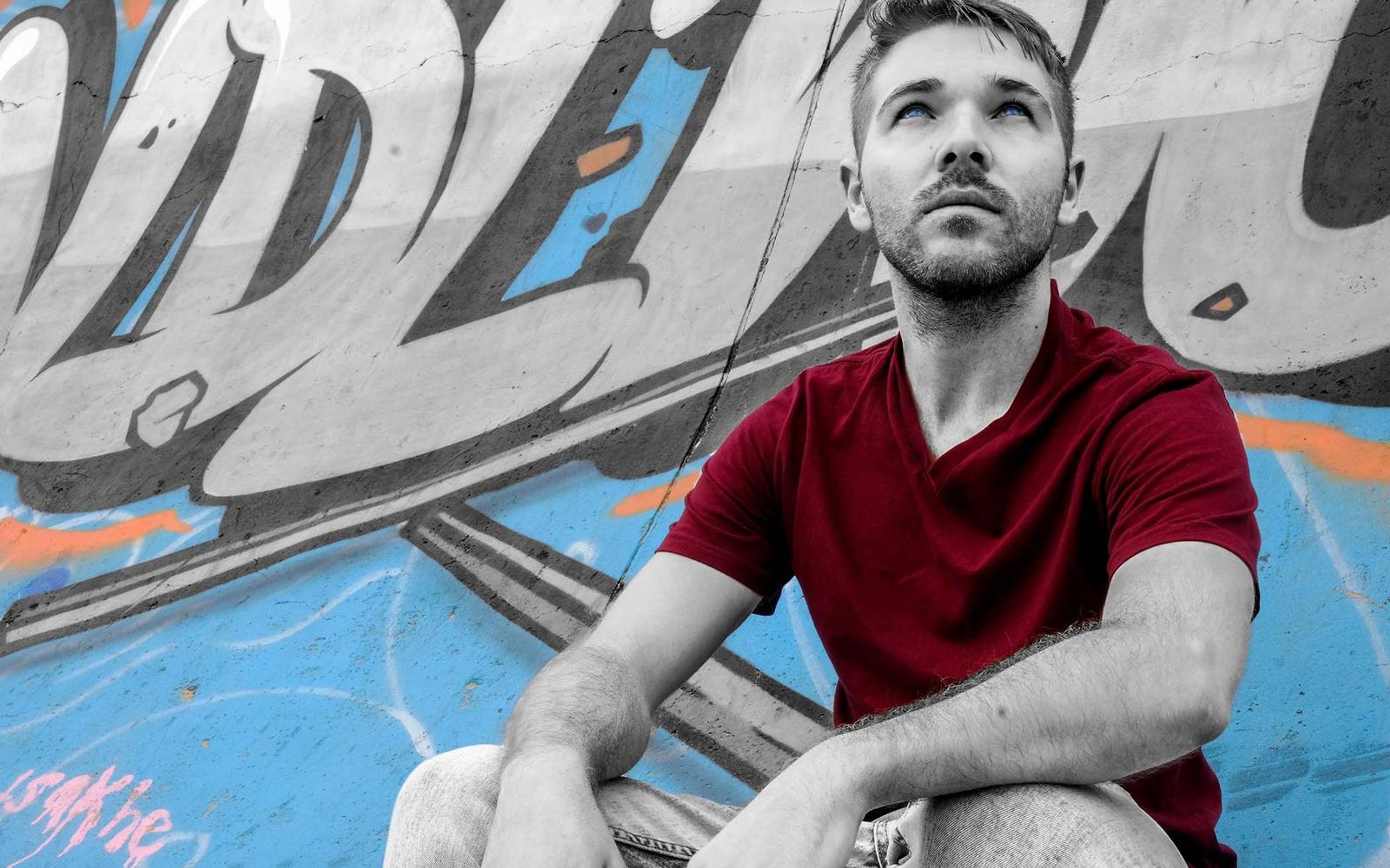 Black and white portrait with the maroon tee kept in colour, man looking upward in front of graffiti lettering