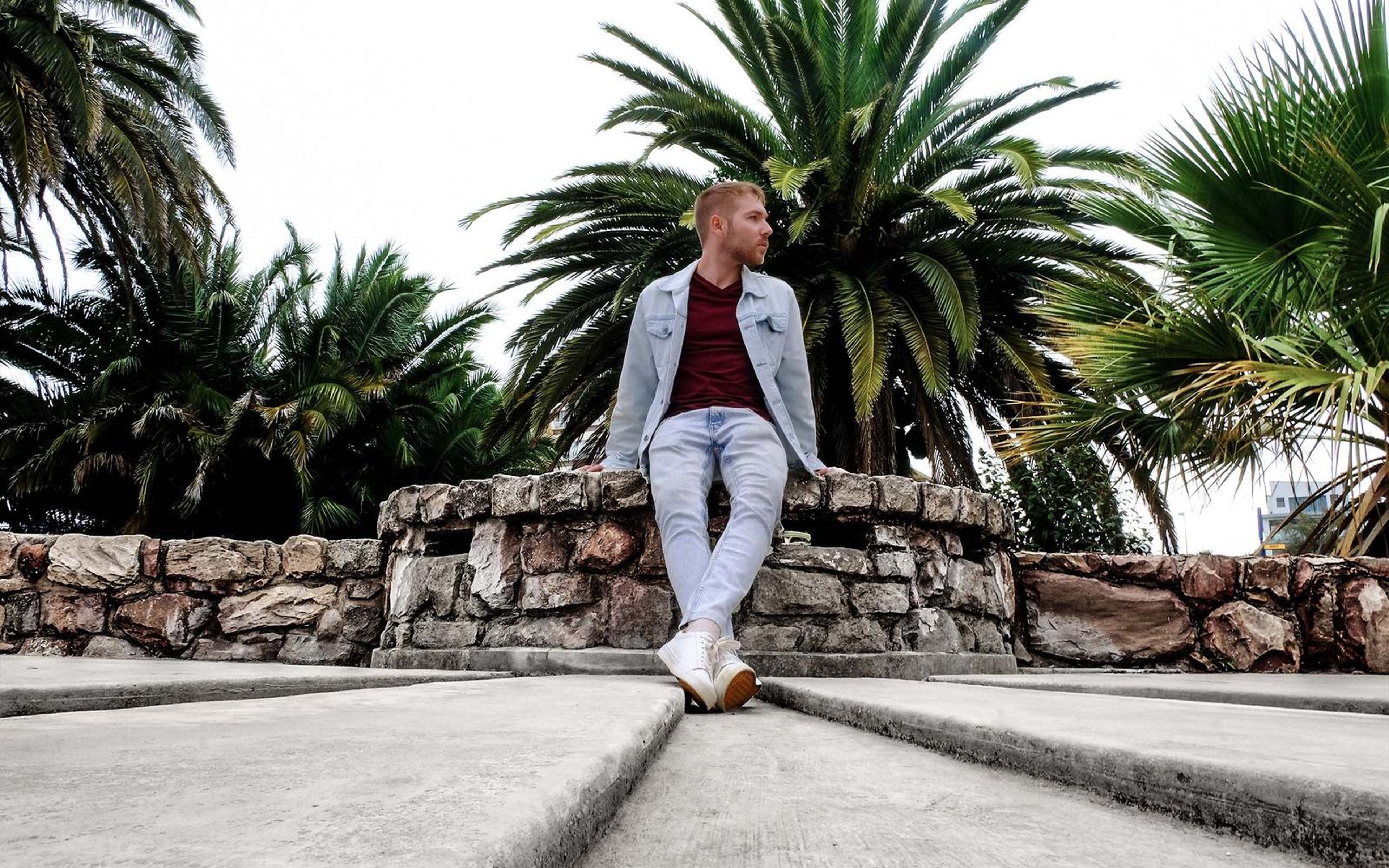Man in a pale jacket seated on a stone wall between palm trees on a cloudy day