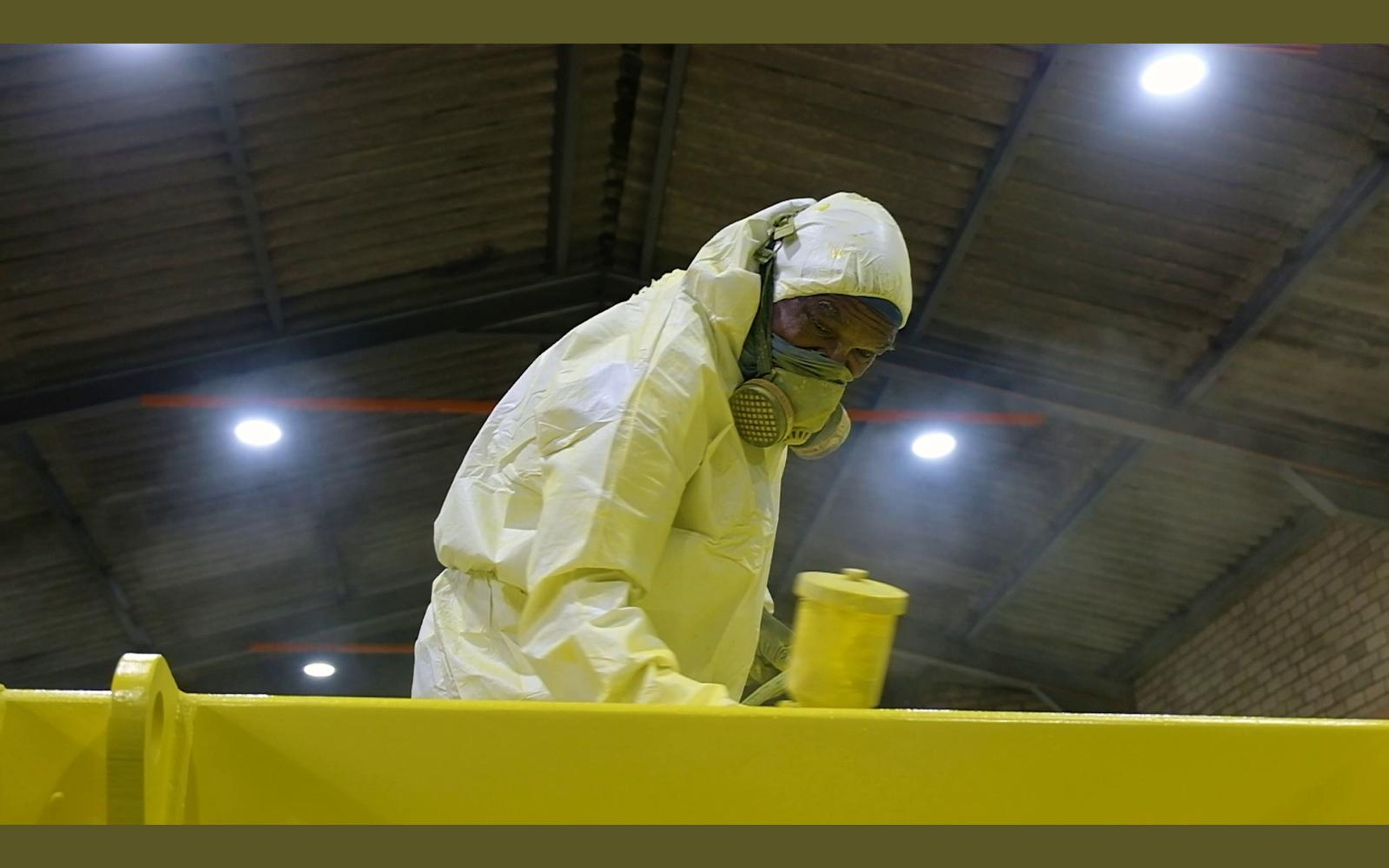 Painter in a white suit and respirator leaning over a yellow steel beam under the roof lights