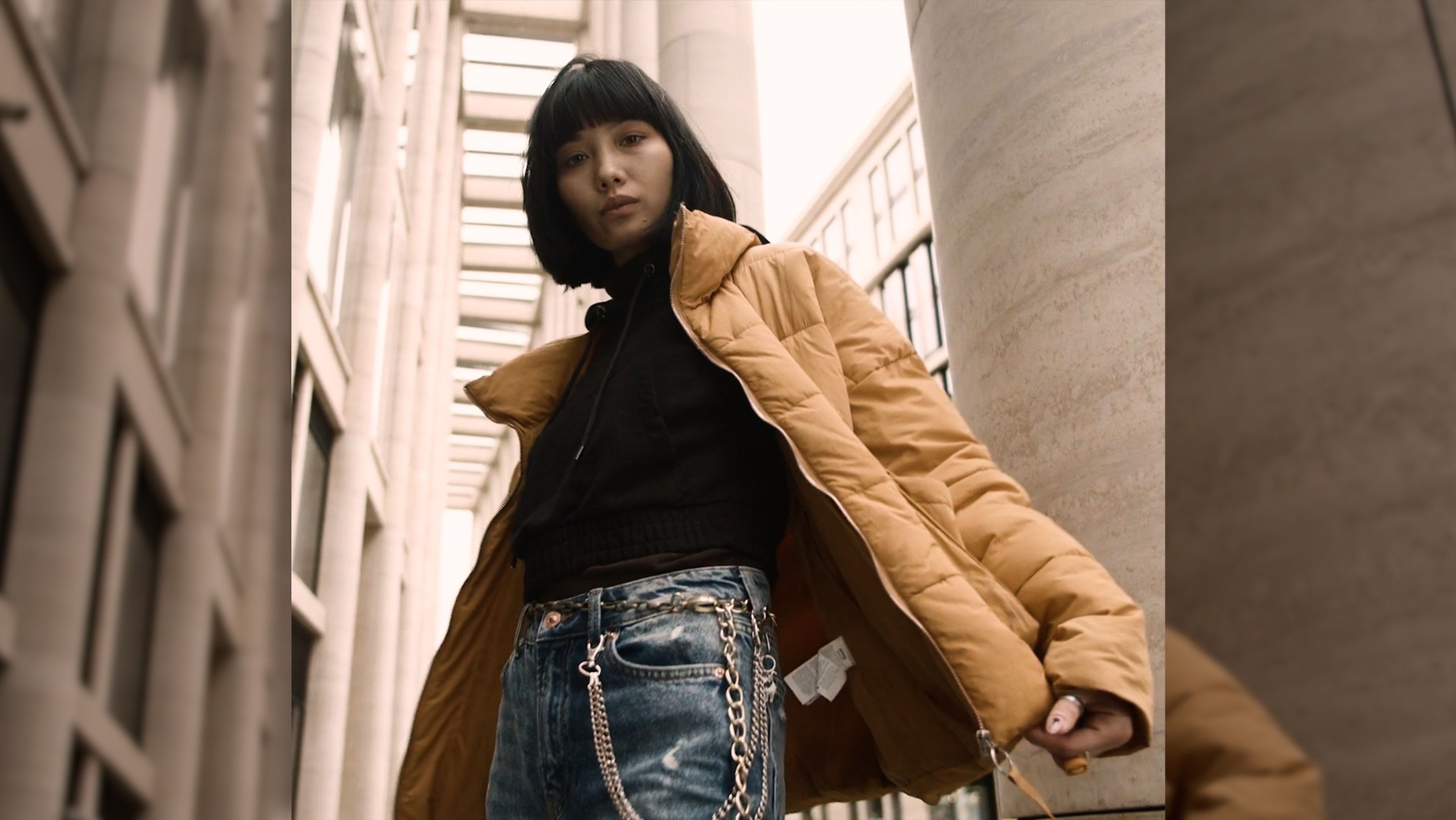 Woman in a tan puffer jacket and jeans posed against tall stone columns