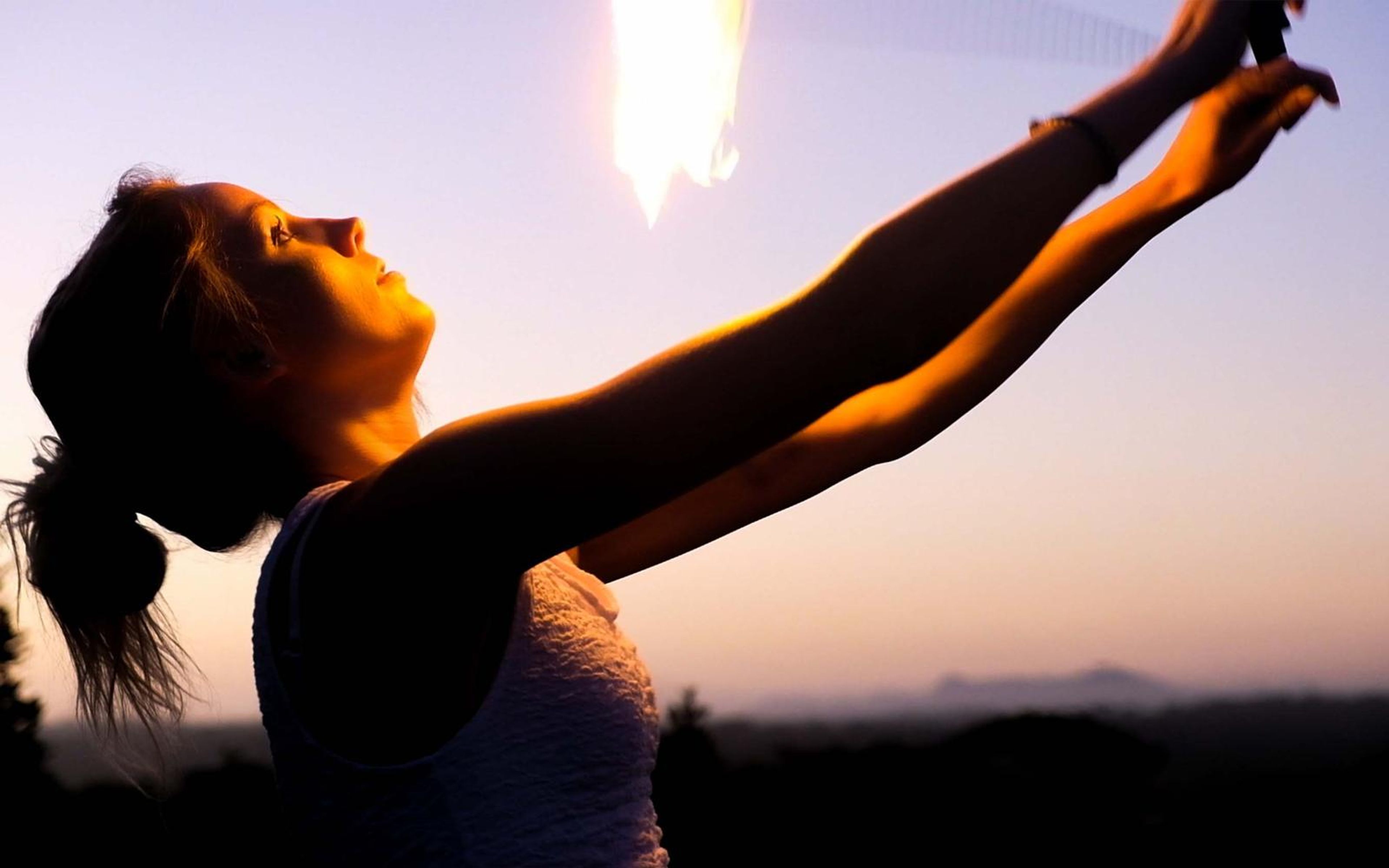 Woman in a white top reaching up with both arms as a fire poi flares above, hills on the horizon