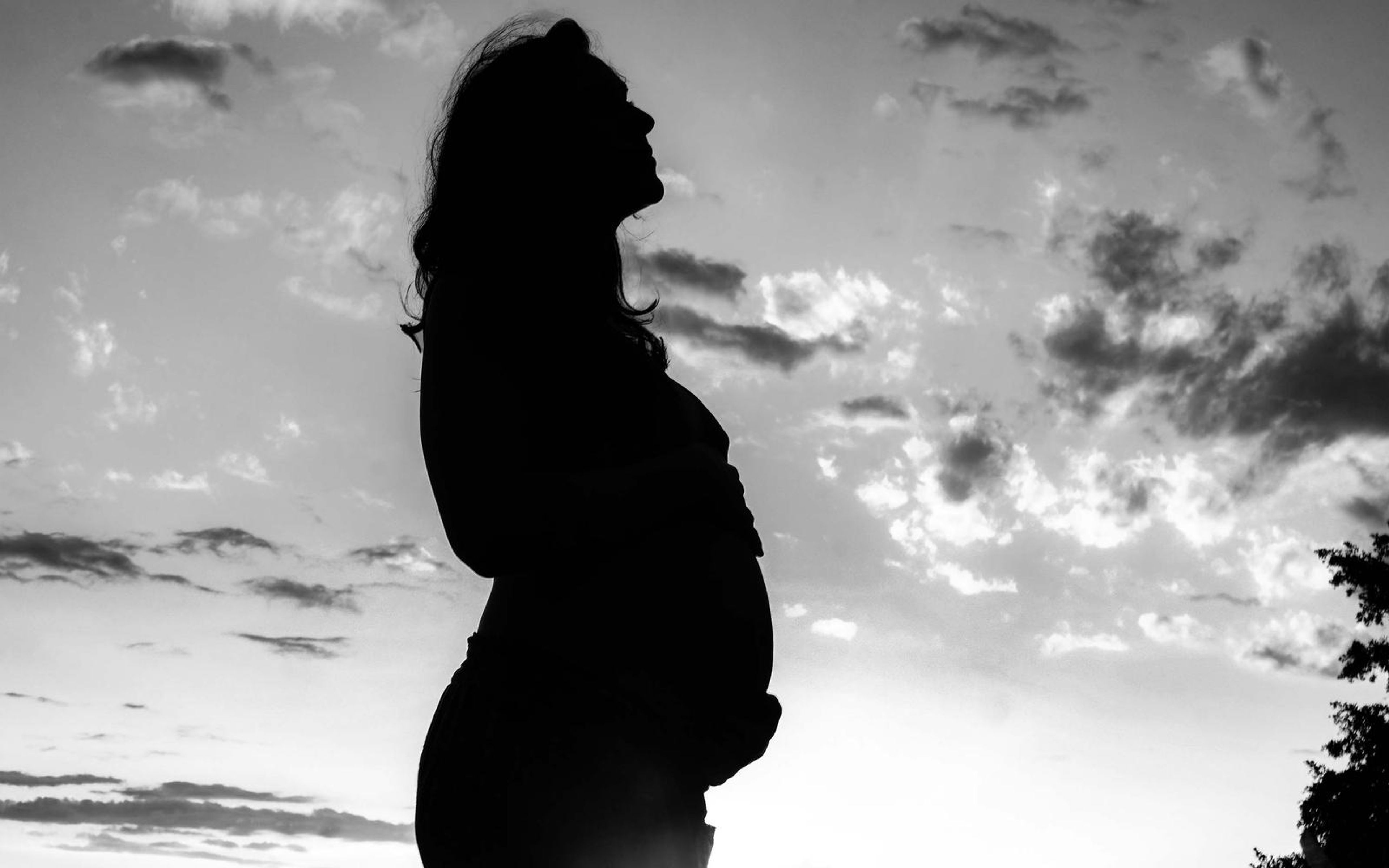 Black and white profile of a pregnant woman looking up, hands on her belly, dramatic clouds