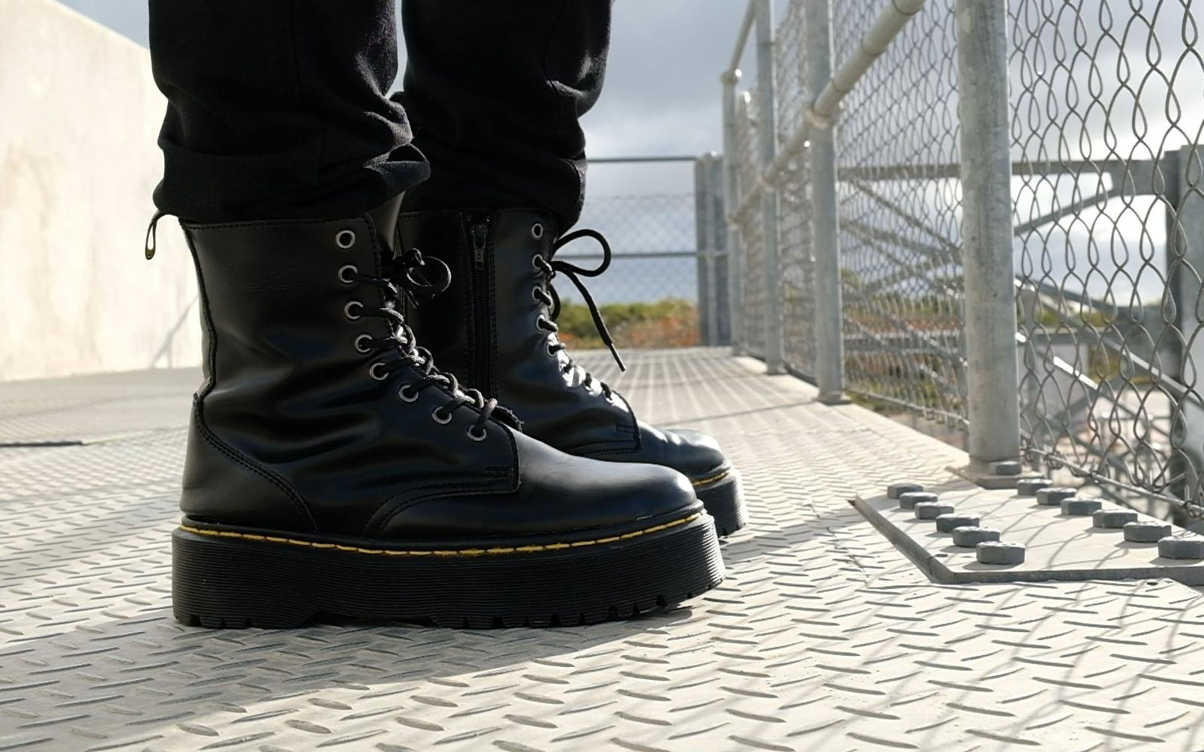 Black lace-up platform boots with yellow stitching standing on a steel plate floor beside a wire fence