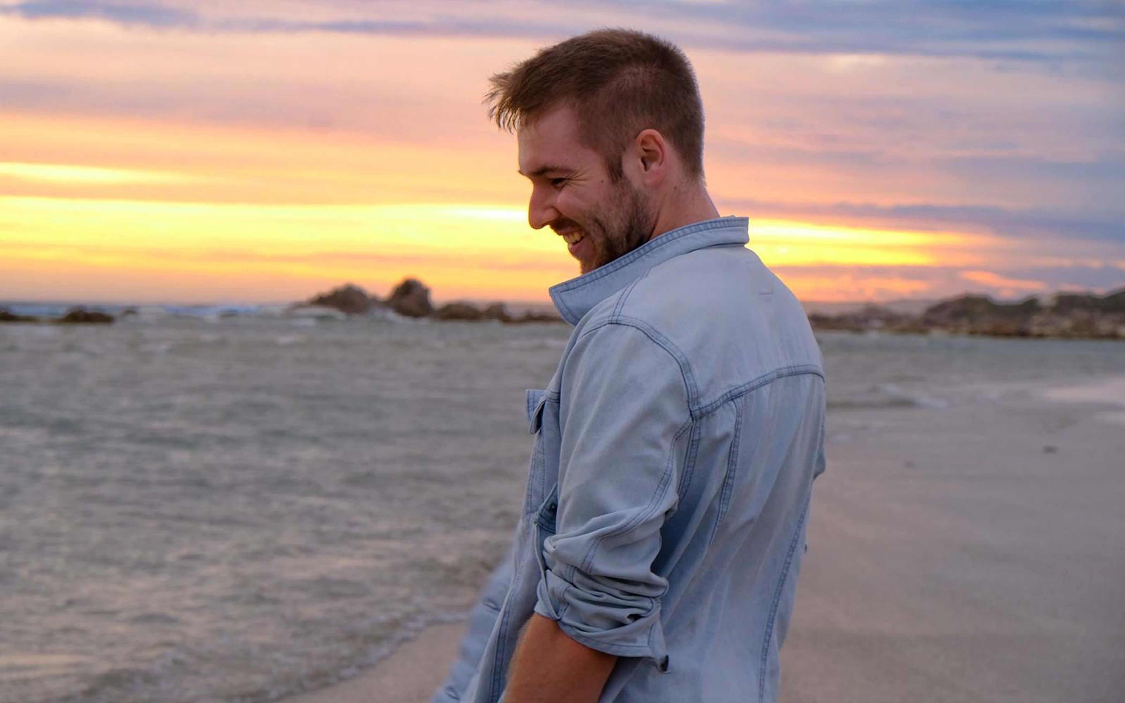 Man smiling and looking down on the beach at sunset, jacket sleeves rolled up