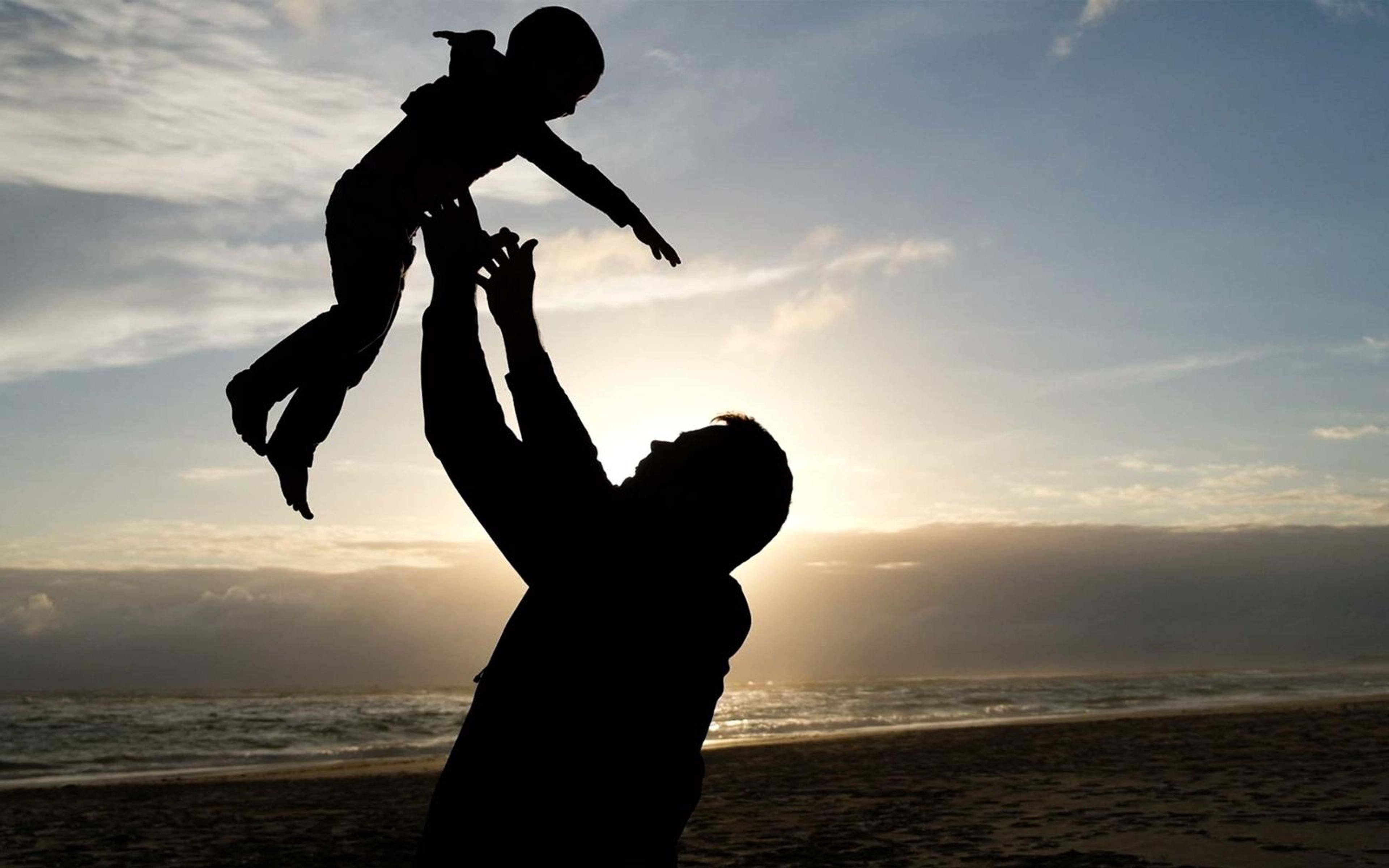 Silhouette of a man lifting a small child into the air against a cloudy sunset sky over a beach