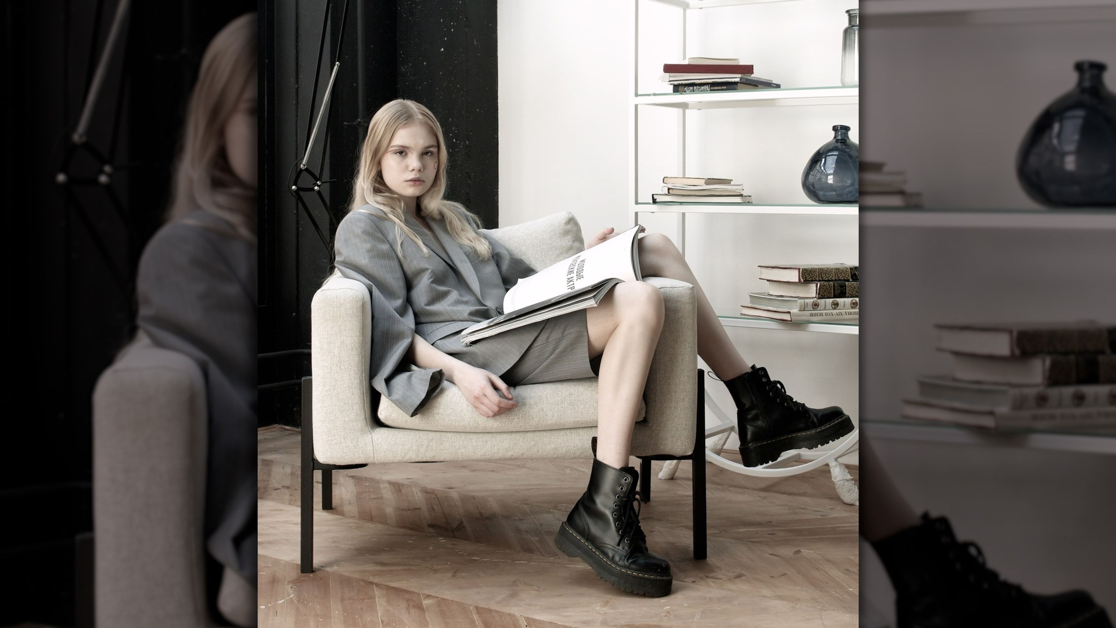 Blonde woman in a grey jacket seated in an armchair with shelves behind her