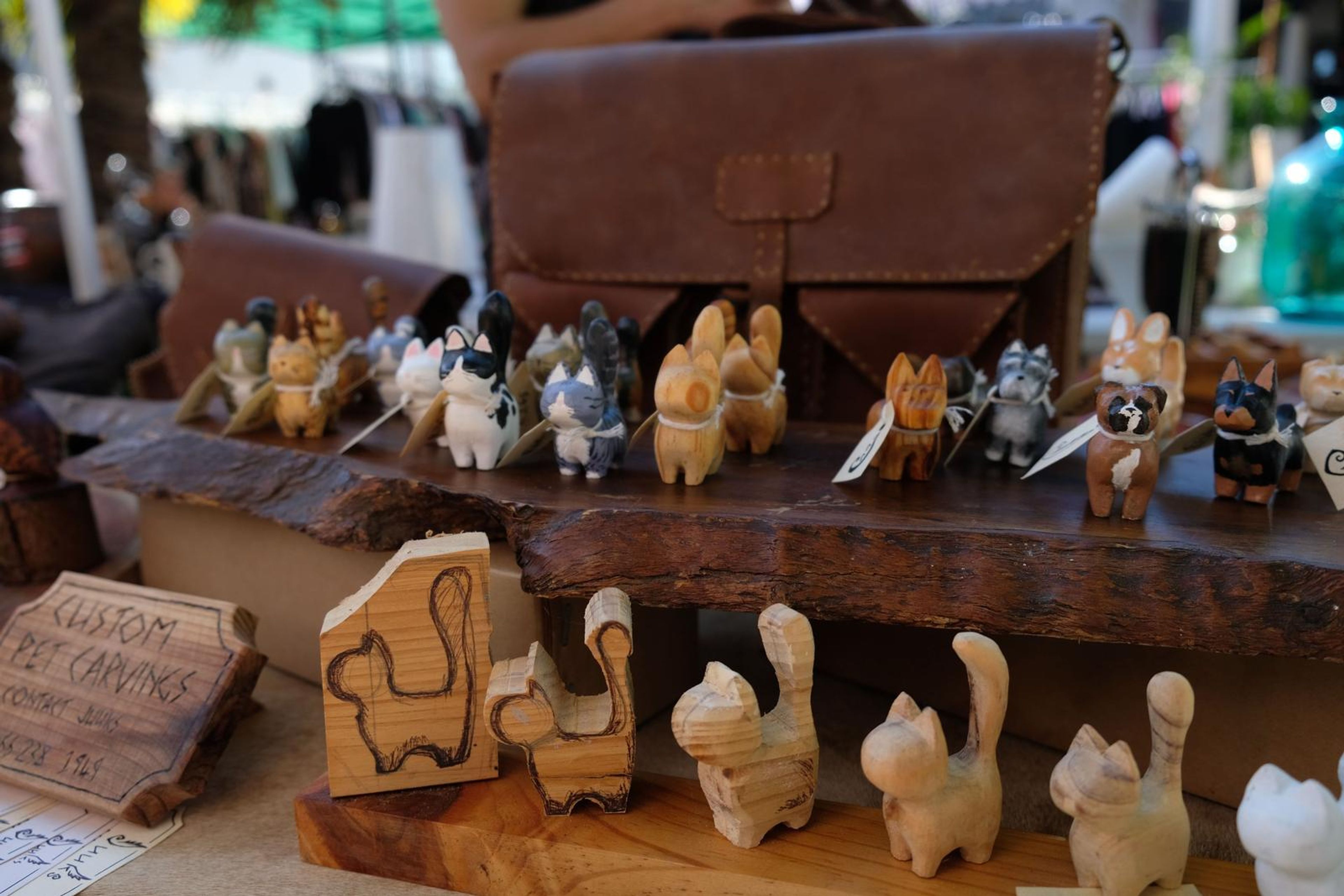 Hand-carved wooden pet figures on a bark slab behind a leather satchel