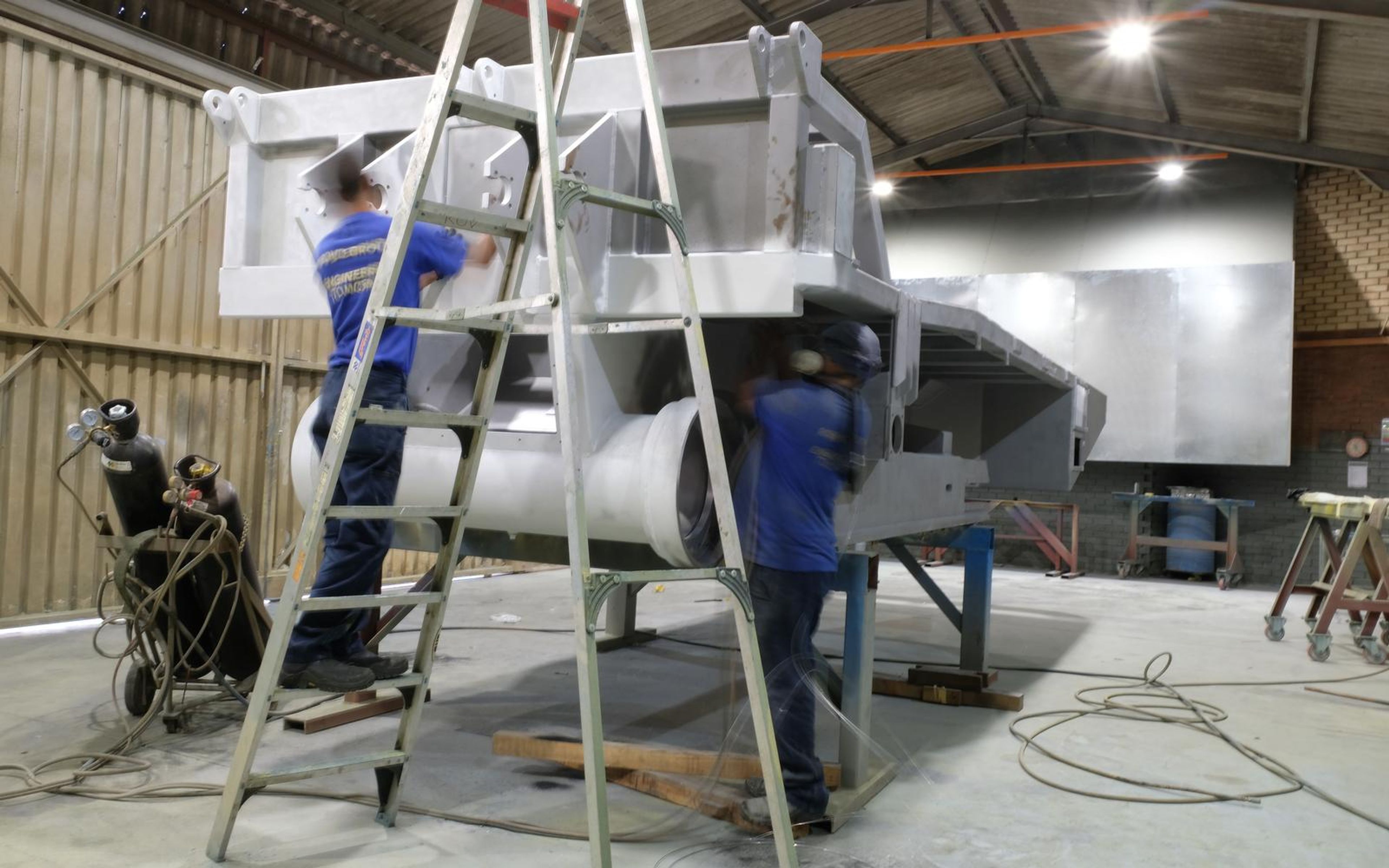 Painters on a ladder spraying a large grey steel component in the paint bay