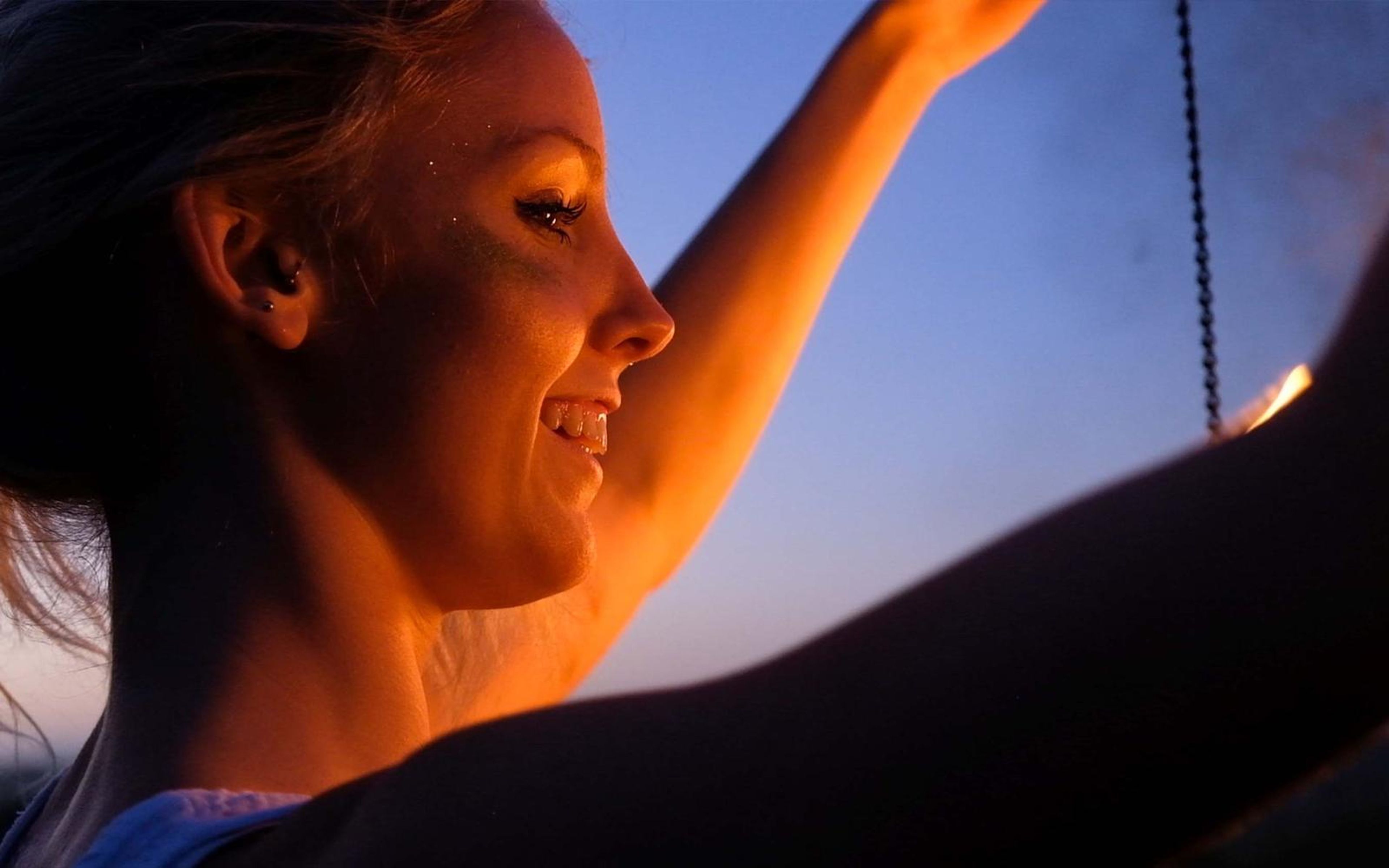 Smiling close-up in profile with one arm raised, blue dusk sky and a hanging chain behind